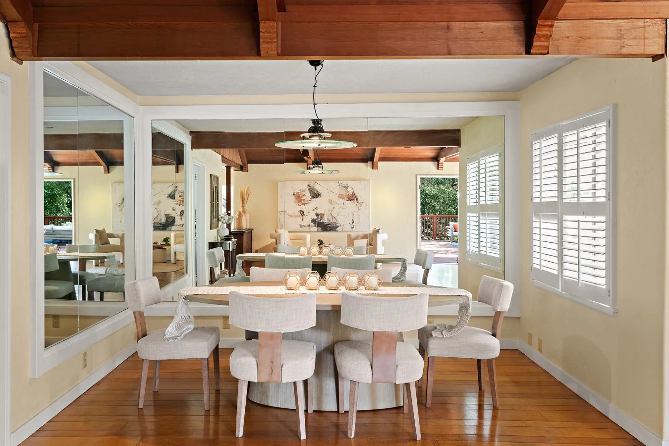 Bright dining room with a large table, upholstered chairs, mirrored wall, white shutters, and exposed wood-beam ceiling