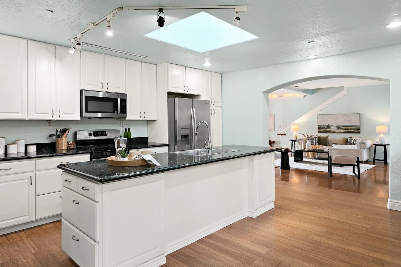 Bright white kitchen with black granite island, stainless steel appliances, and an arched opening to a furnished living room