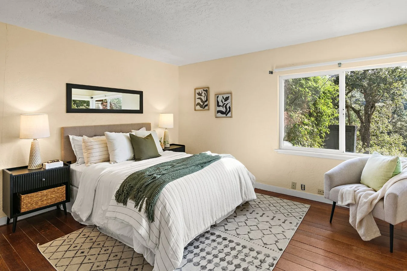 Bright contemporary bedroom with a neatly made bed, green throw blanket, patterned rug, armchair, and large window overlooking trees