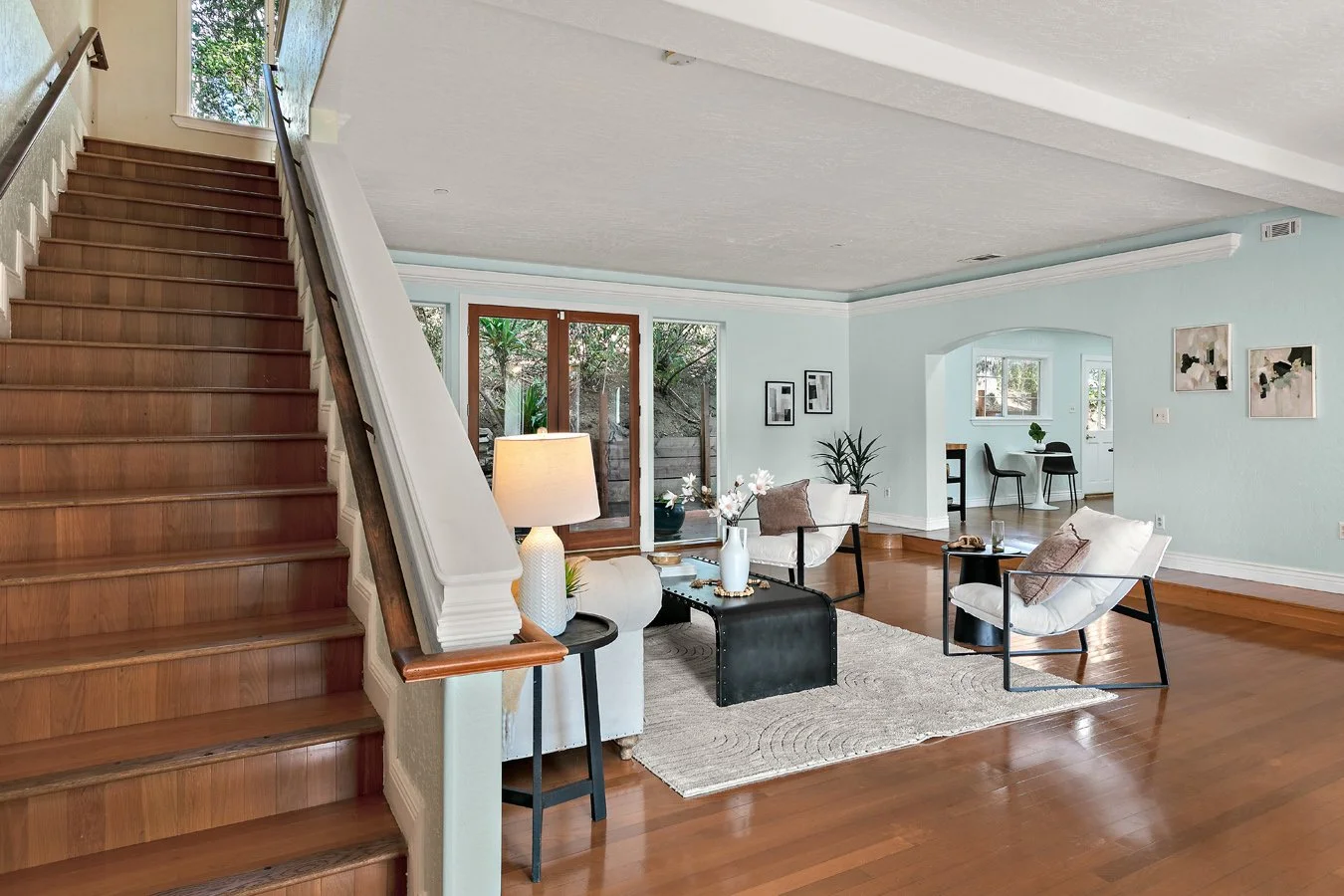 Bright open-concept living room with hardwood floors, white seating, black coffee table, pale blue walls, and staircase