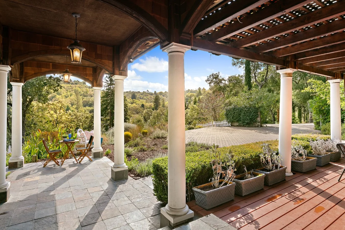 Covered outdoor patio with white columns, wooden beams, potted plants, seating, and views of a wooded hillside