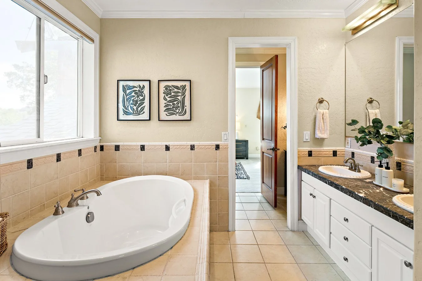 Bright beige-tiled bathroom with a large corner soaking tub, double vanity, mirrored wall, window, floral artwork, and doorway.