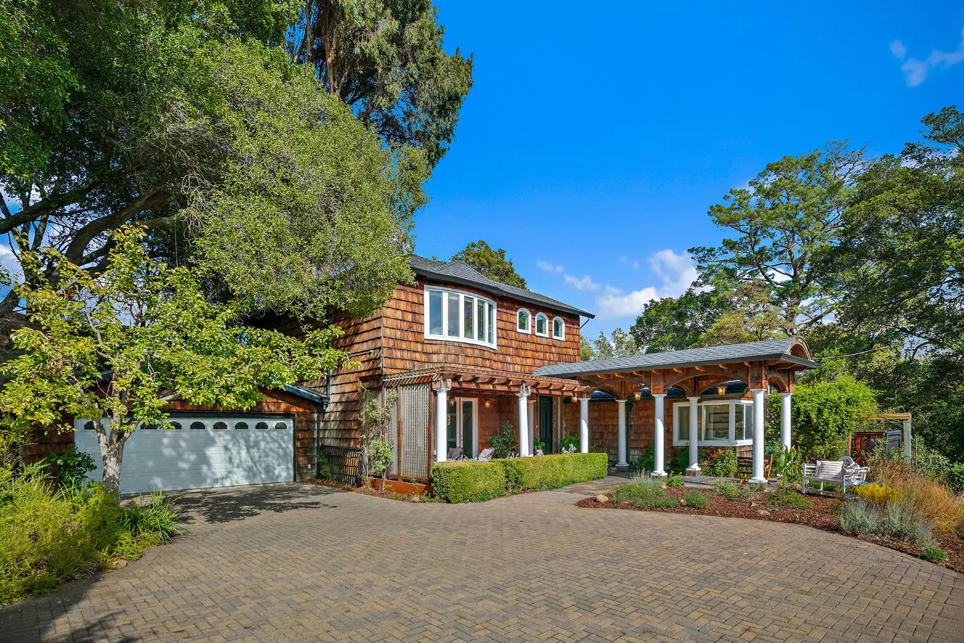 Wood-shingled suburban home with covered porch, attached garage, landscaped yard, and brick driveway under a clear blue sky