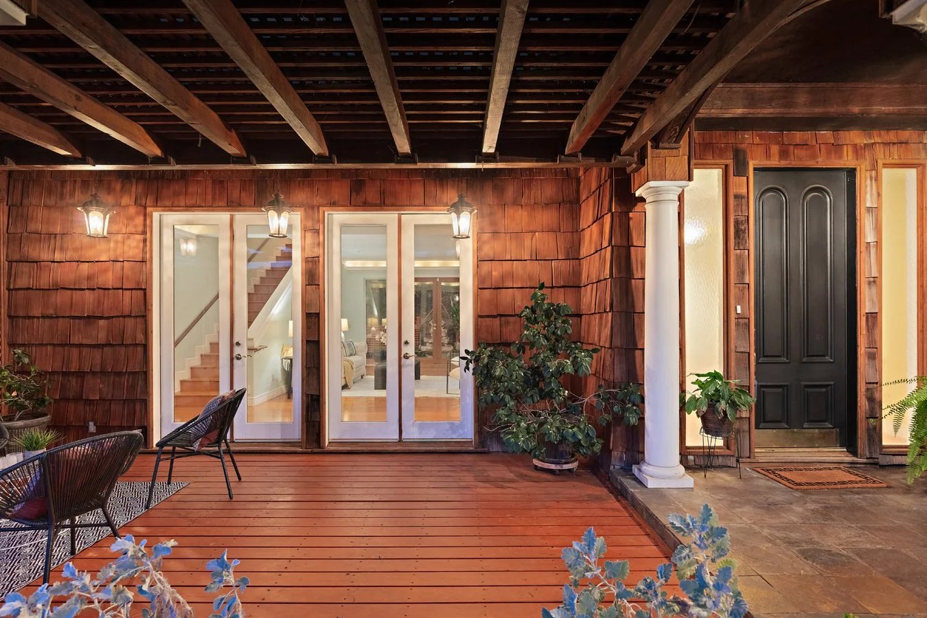 Covered wood-shingled front porch with exposed beams, glass double doors, black entry door, white column, hanging lanterns, and potted plants