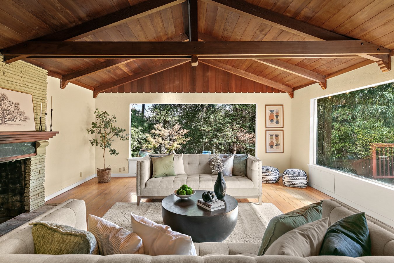 Spacious midcentury modern living room with exposed wood-beam ceiling, fireplace, sofas, round coffee table, and large windows overlooking trees