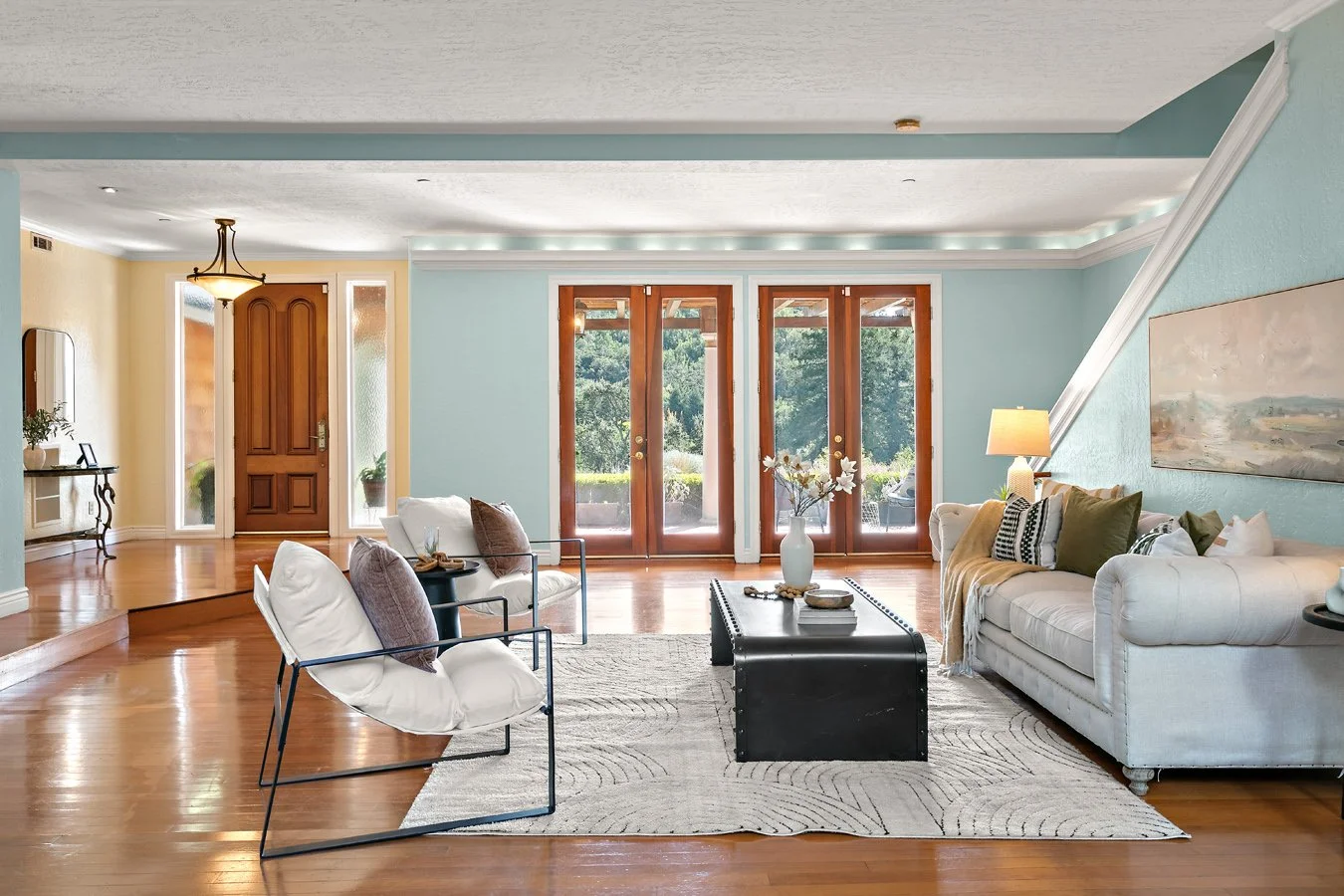 Bright blue living room with hardwood floors, white sofas, lounge chairs, coffee table, wall art, and French doors overlooking a garden.