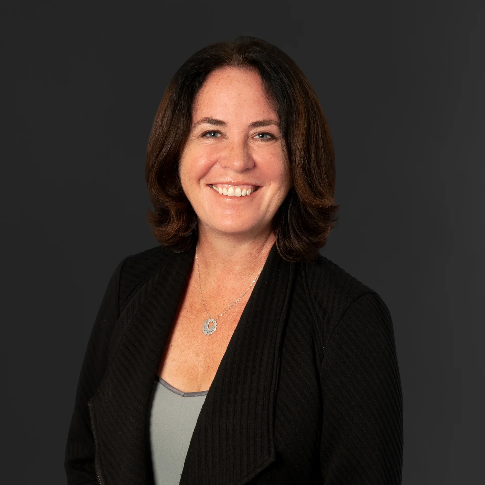 Professional headshot of a smiling woman with shoulder-length dark hair wearing a black cardigan and floral pendant necklace