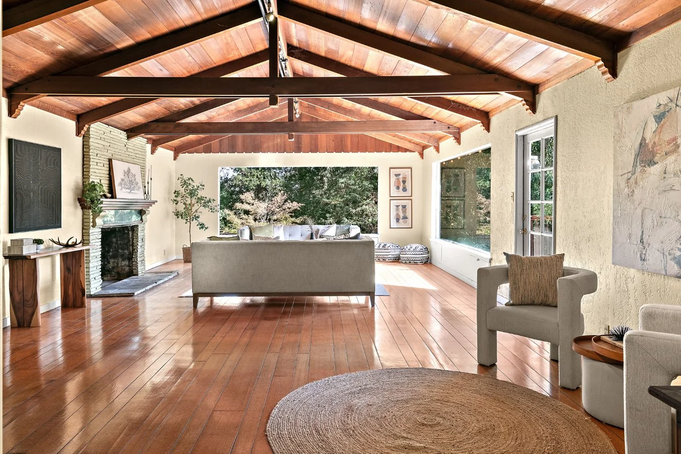 Bright midcentury living room with exposed wood-beam ceiling, fireplace, hardwood floors, sofa, armchairs, and large garden-view windows