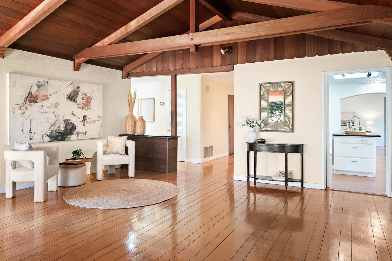 Bright open-concept living room with exposed wood-beam ceiling, hardwood floors, white armchairs, abstract artwork, console table, and kitchen beyond