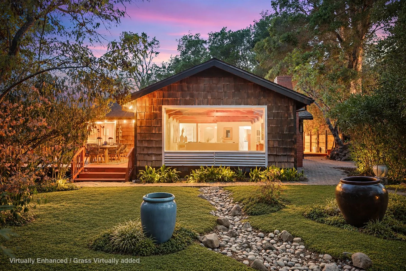 Warmly lit shingle-clad cottage with open living room, covered deck, landscaped lawn, river-rock garden path, and trees at sunset