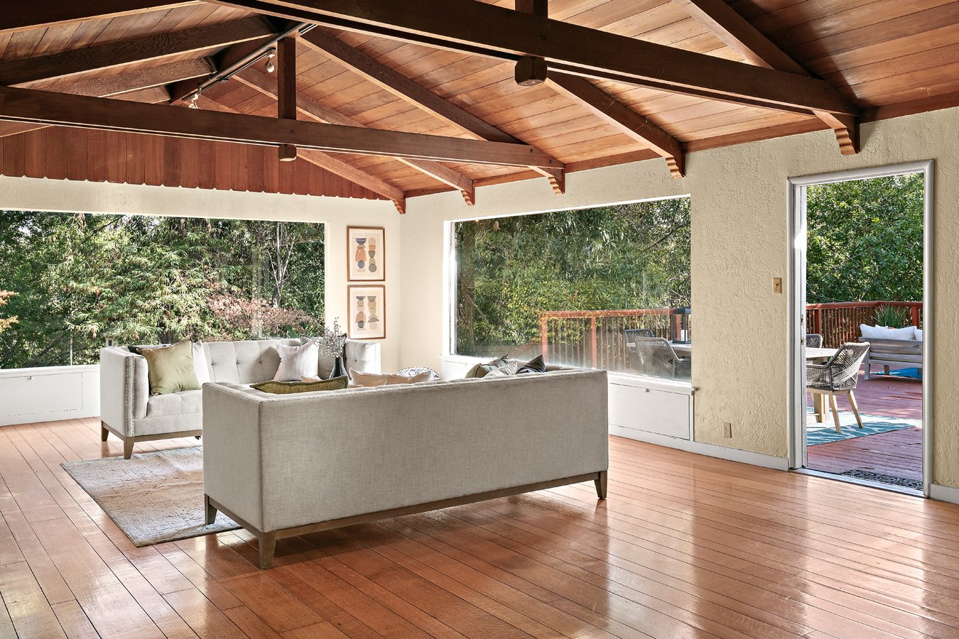 Bright midcentury living room with white sofas, hardwood floors, exposed wood-beam ceiling, and glass doors opening to a deck surrounded by trees