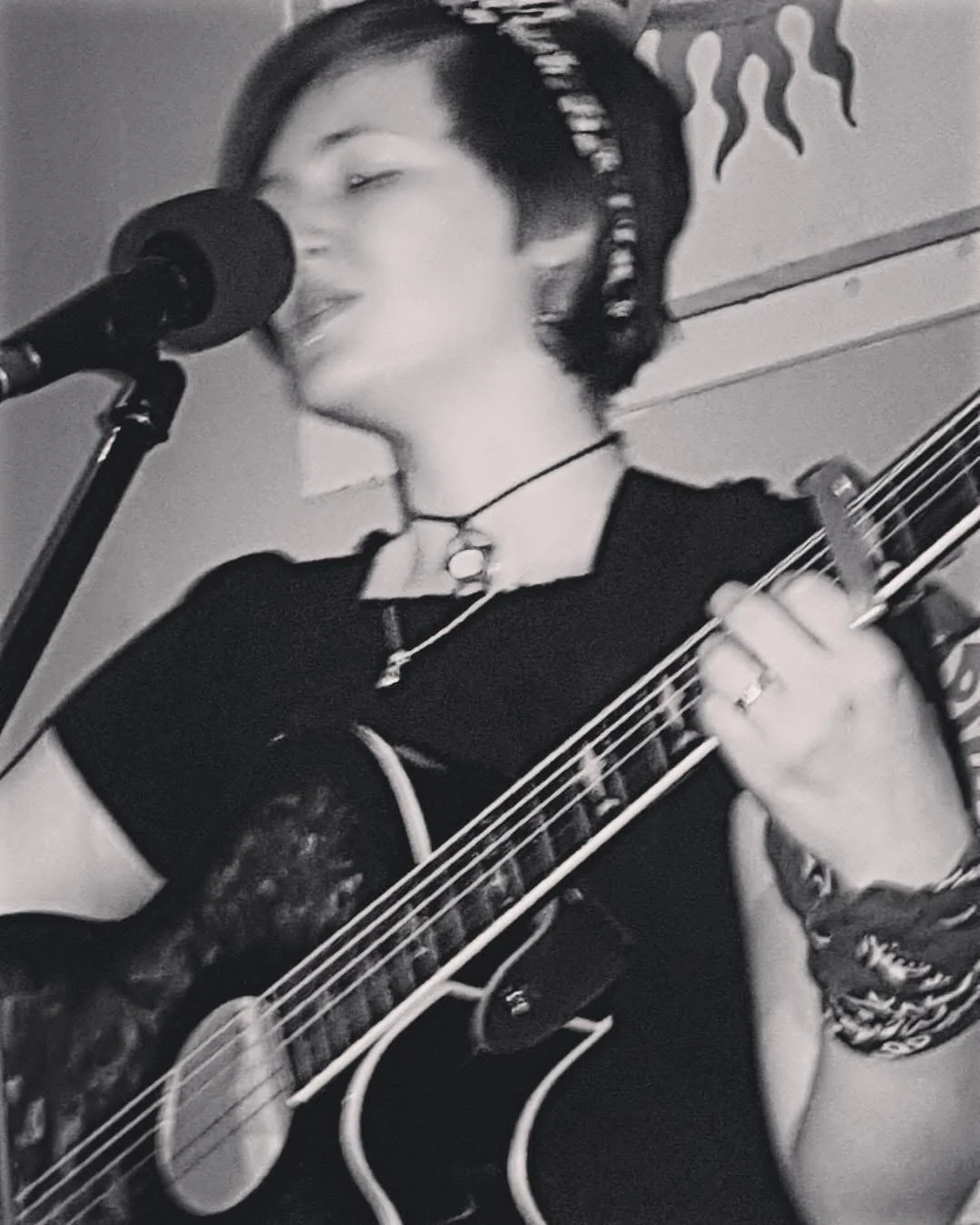 An old photo of Dana Kelly with a pixie hair cut playing the guitar and singing at a gig.
