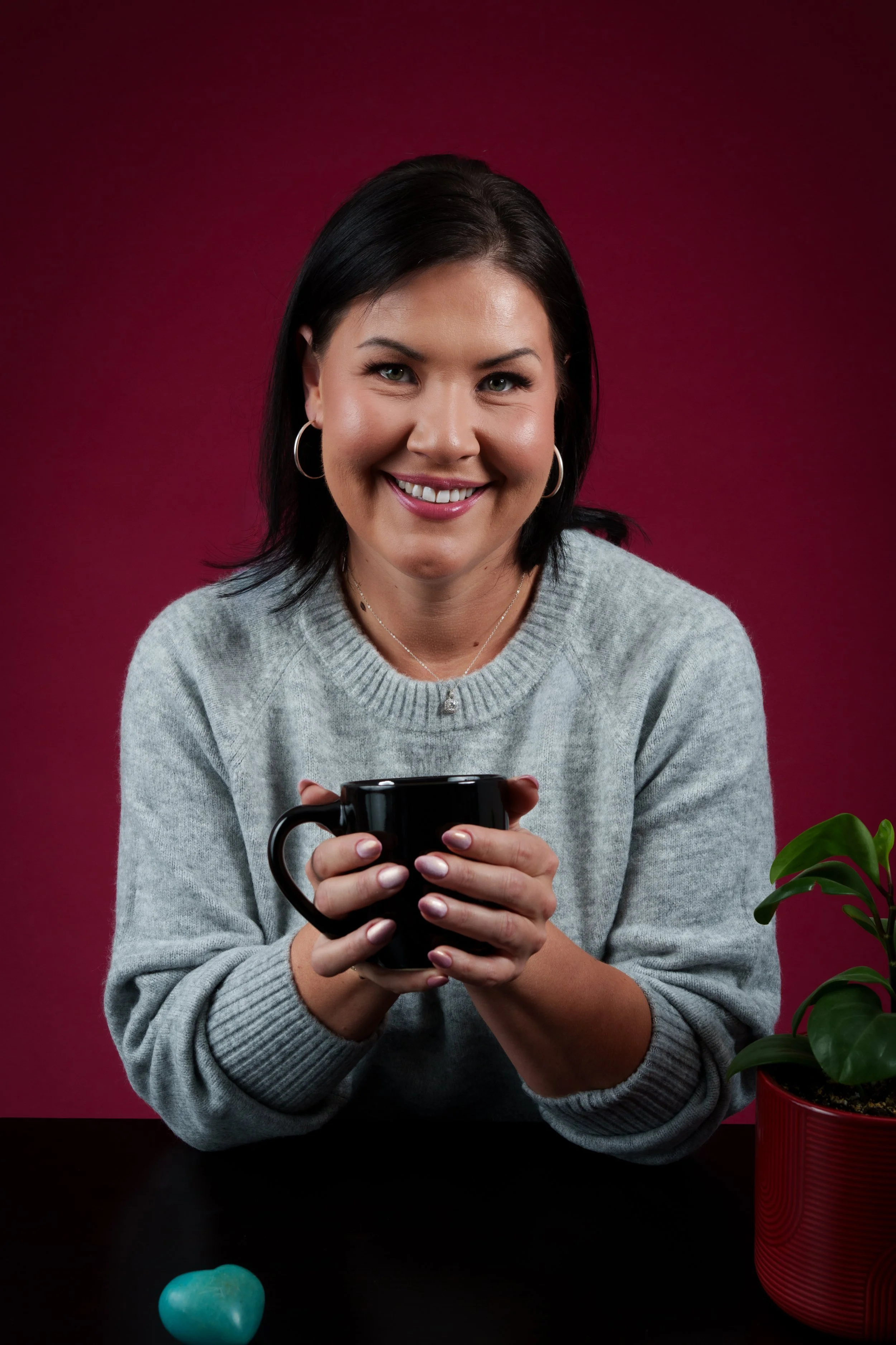 Smiling woman in a gray sweater holding a black mug against a burgundy background