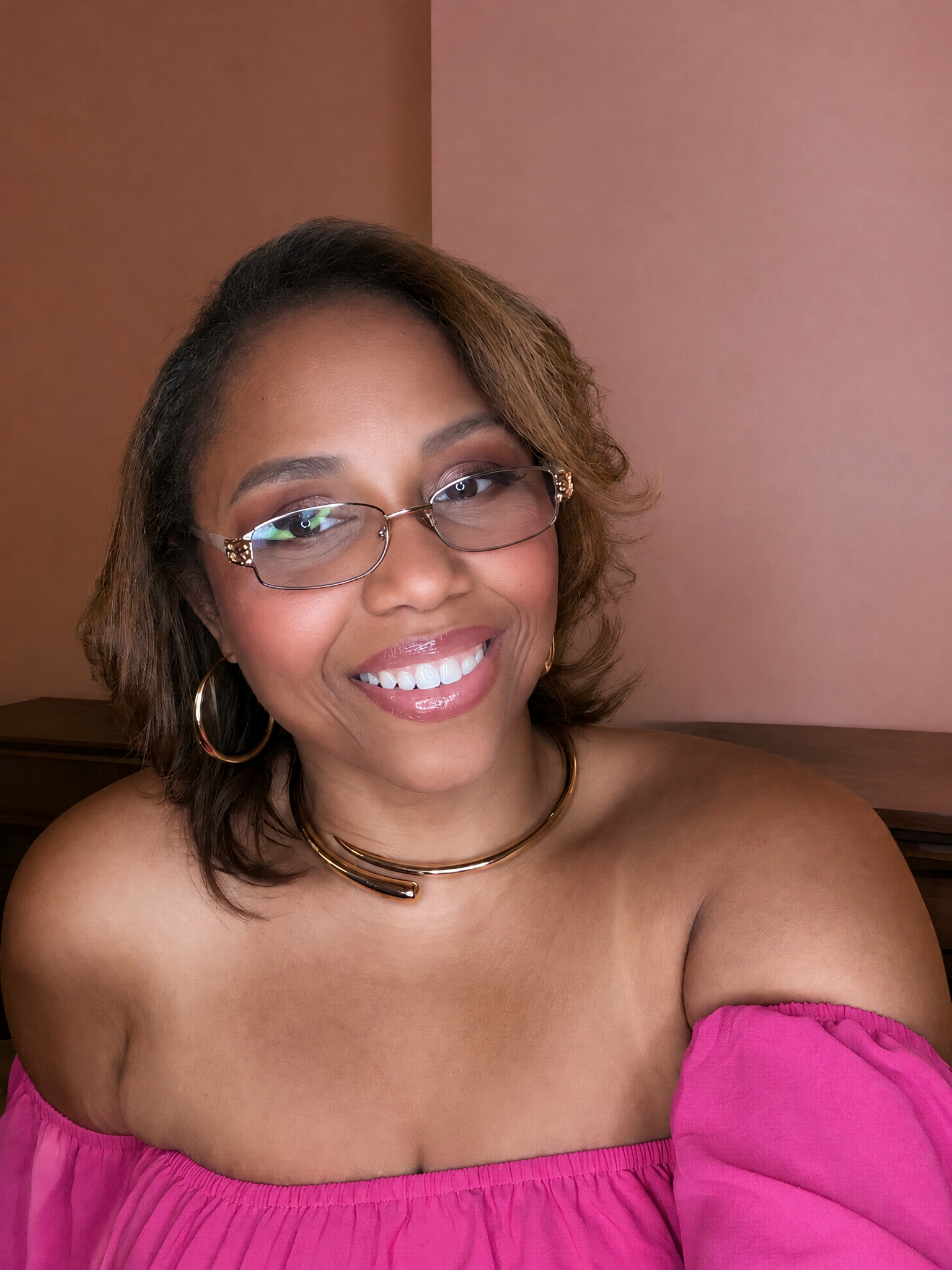 Smiling woman wearing glasses, gold hoop earrings, a statement necklace, and a bright pink off-the-shoulder top.