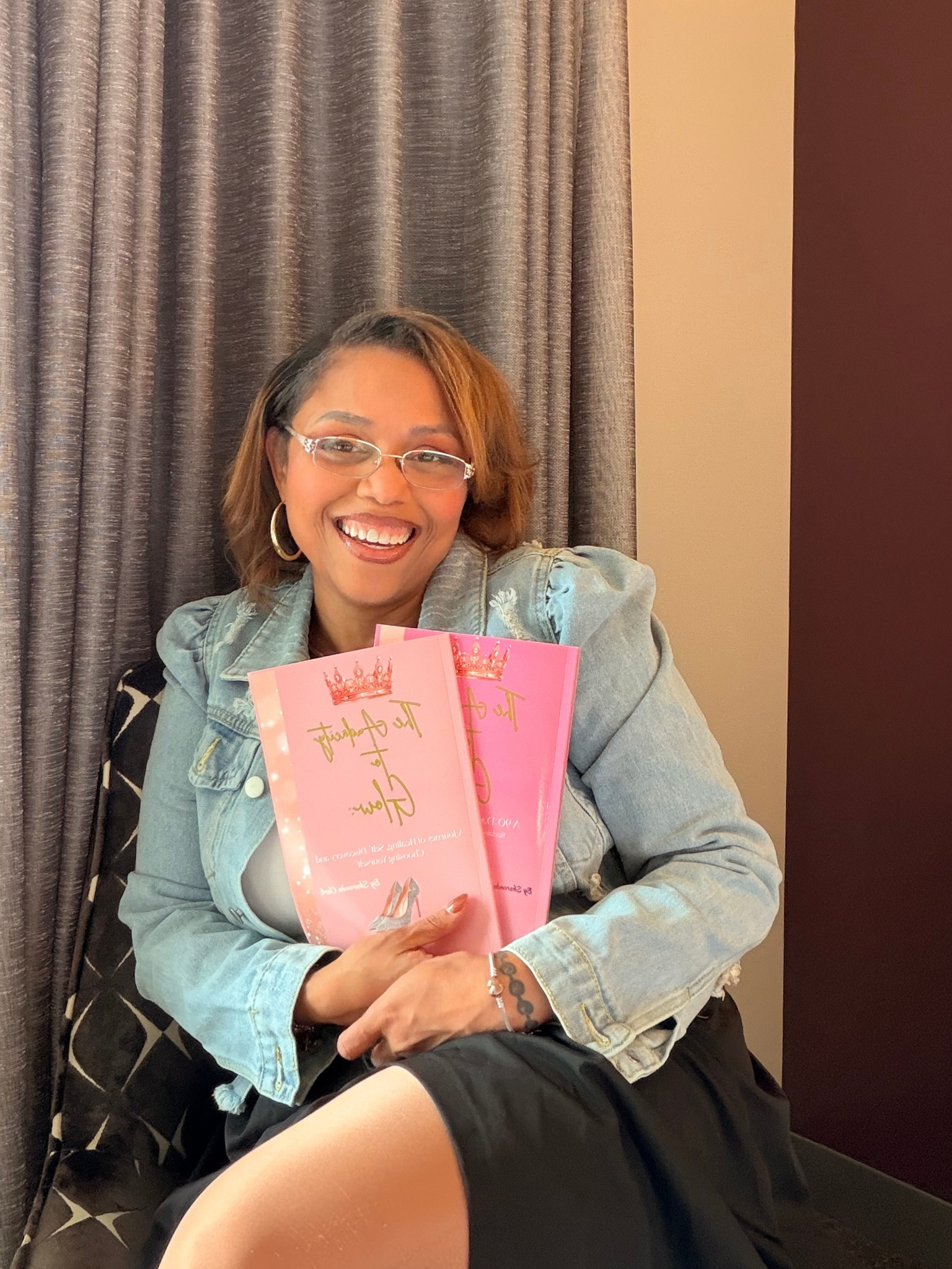 Smiling woman wearing glasses and a denim jacket holds two pink crown-themed books while sitting indoors.