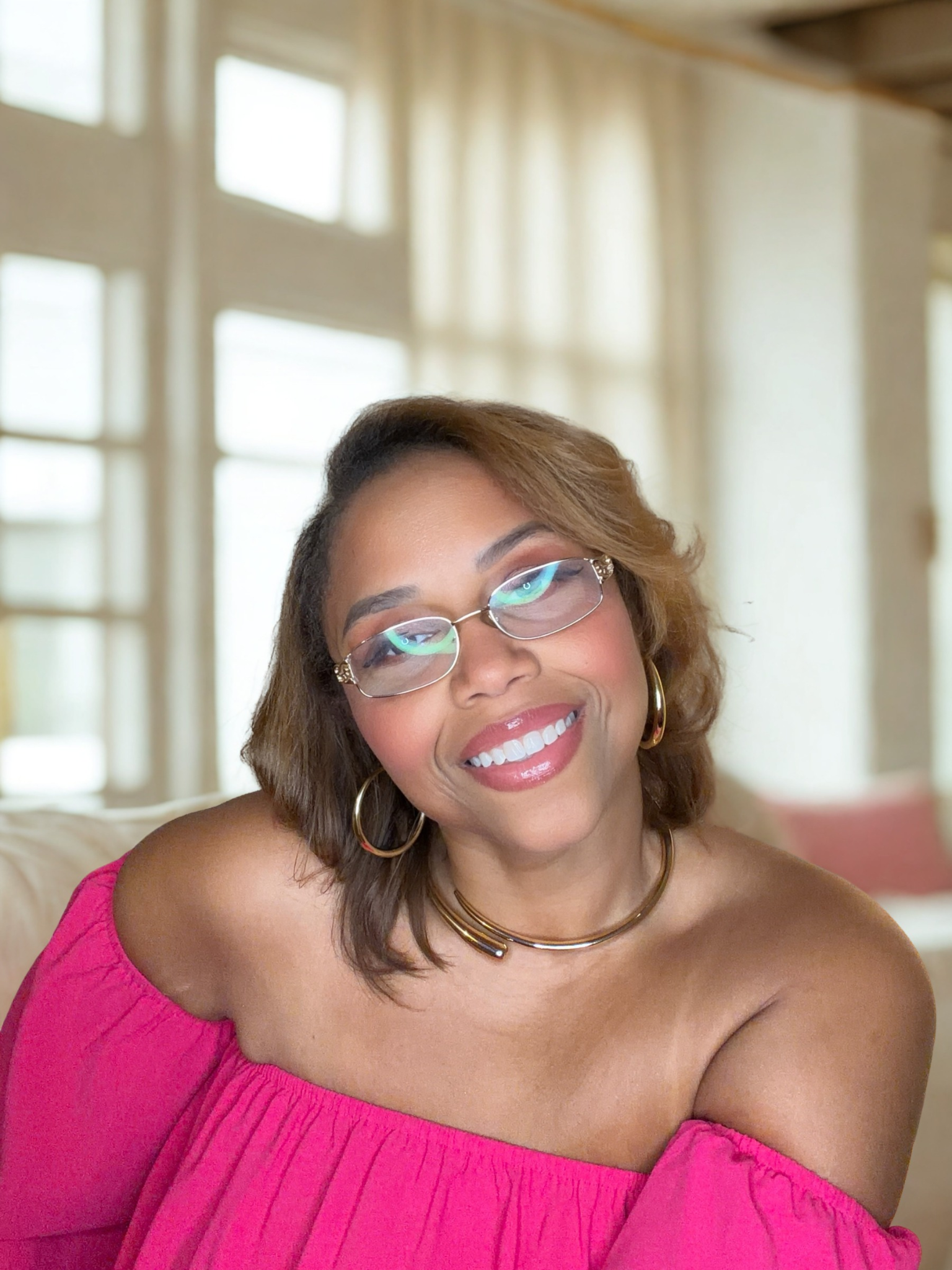 Smiling woman wearing glasses, gold hoop earrings, and a bright pink off-the-shoulder top indoors