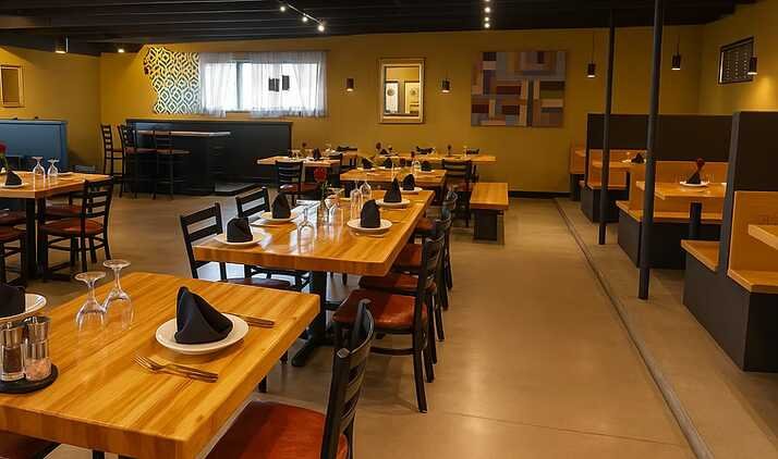 Warmly lit modern restaurant dining room with wooden tables, black napkins, set place settings, and booth seating.
