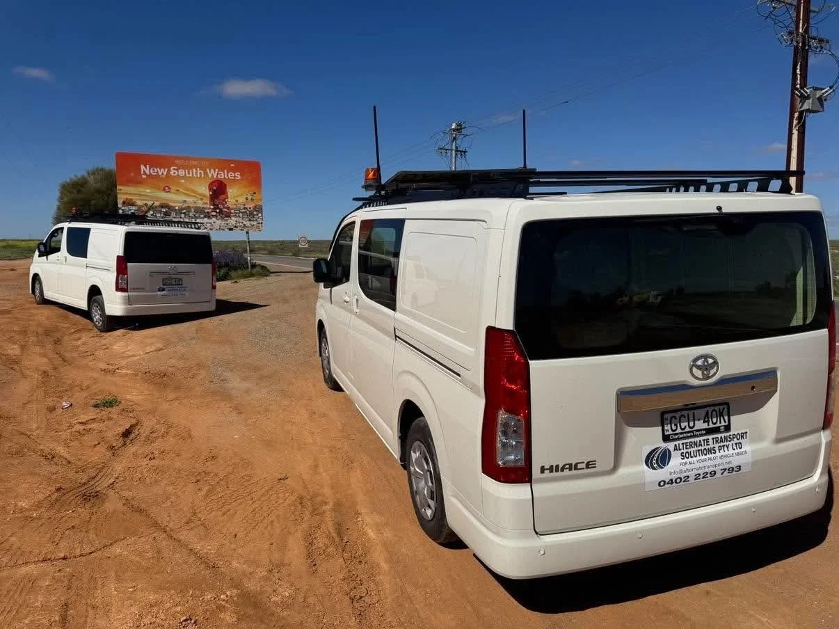 Two white Toyota HiAce vans parked on a dirt roadside near a New South Wales sign.