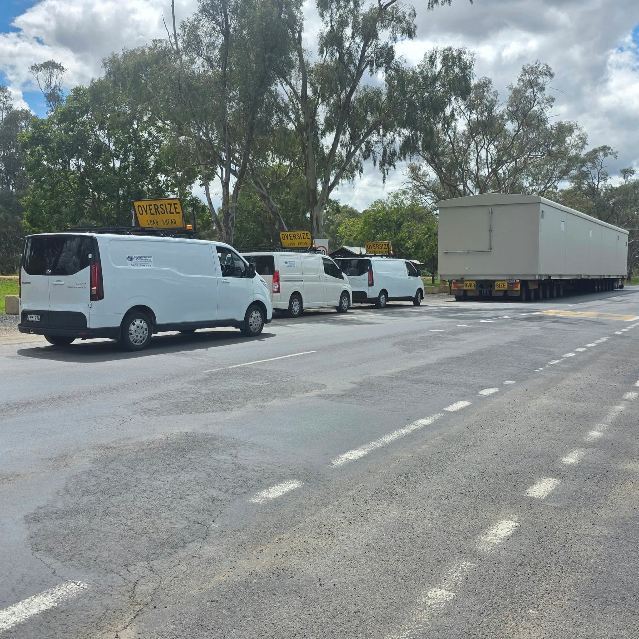 White vans with oversized-load signs escorting a large box trailer on a tree-lined road