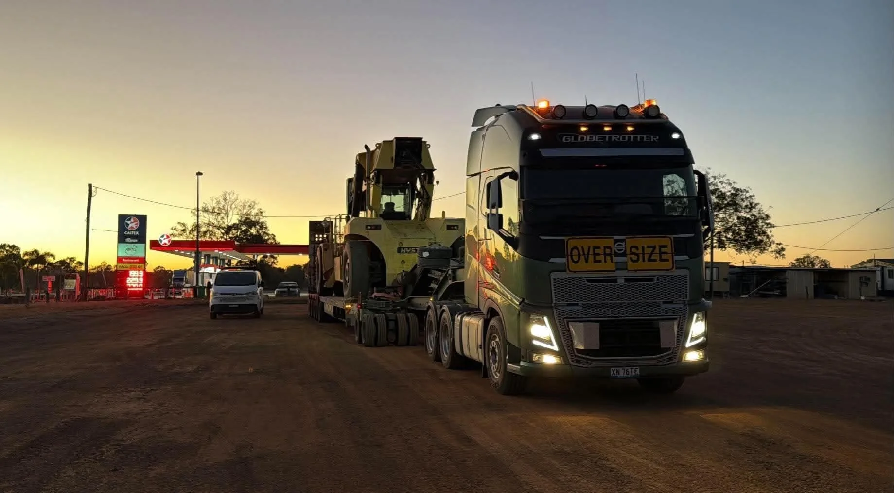 Oversize-load semi-truck hauling heavy construction equipment past a gas station at sunset