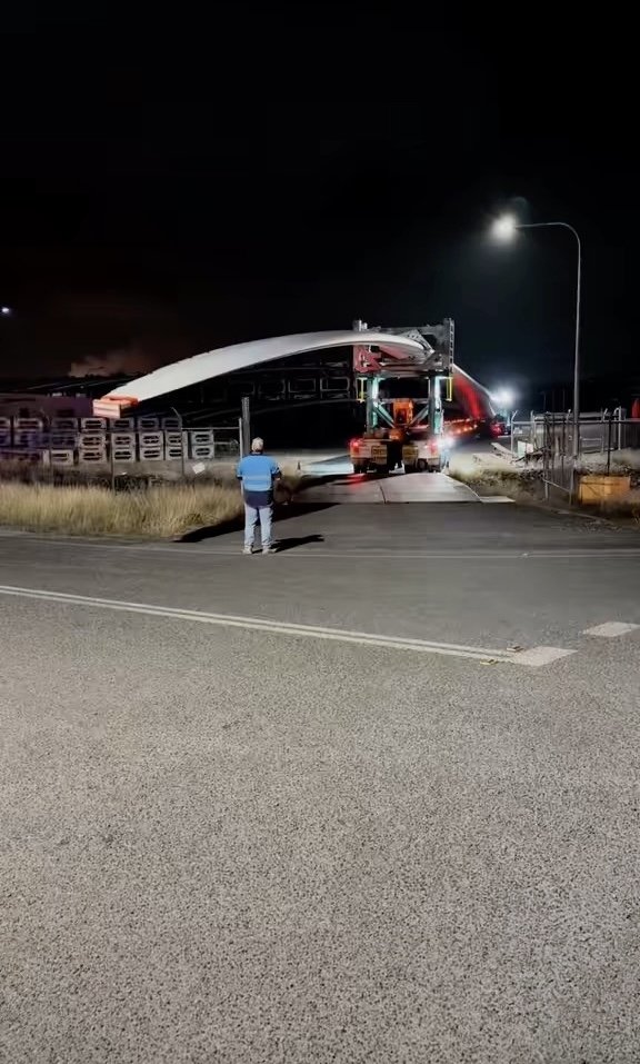 Nighttime view of a large truck hauling an oversized curved structure through an industrial yard, with a person directing traffic.