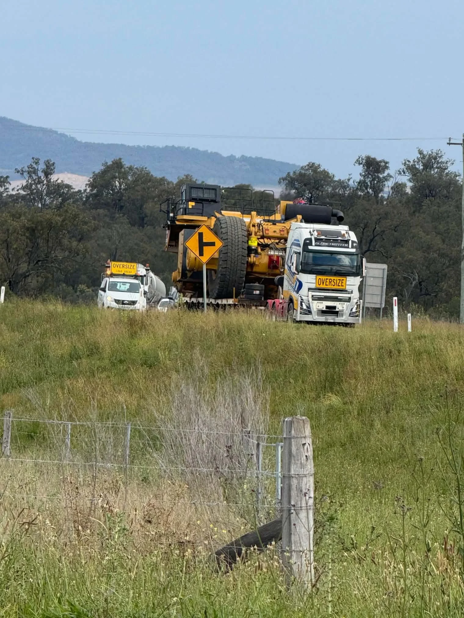 Oversize truck hauling heavy construction equipment on a rural road, with a right-turn warning sign, grassy fields, trees, and mountains.