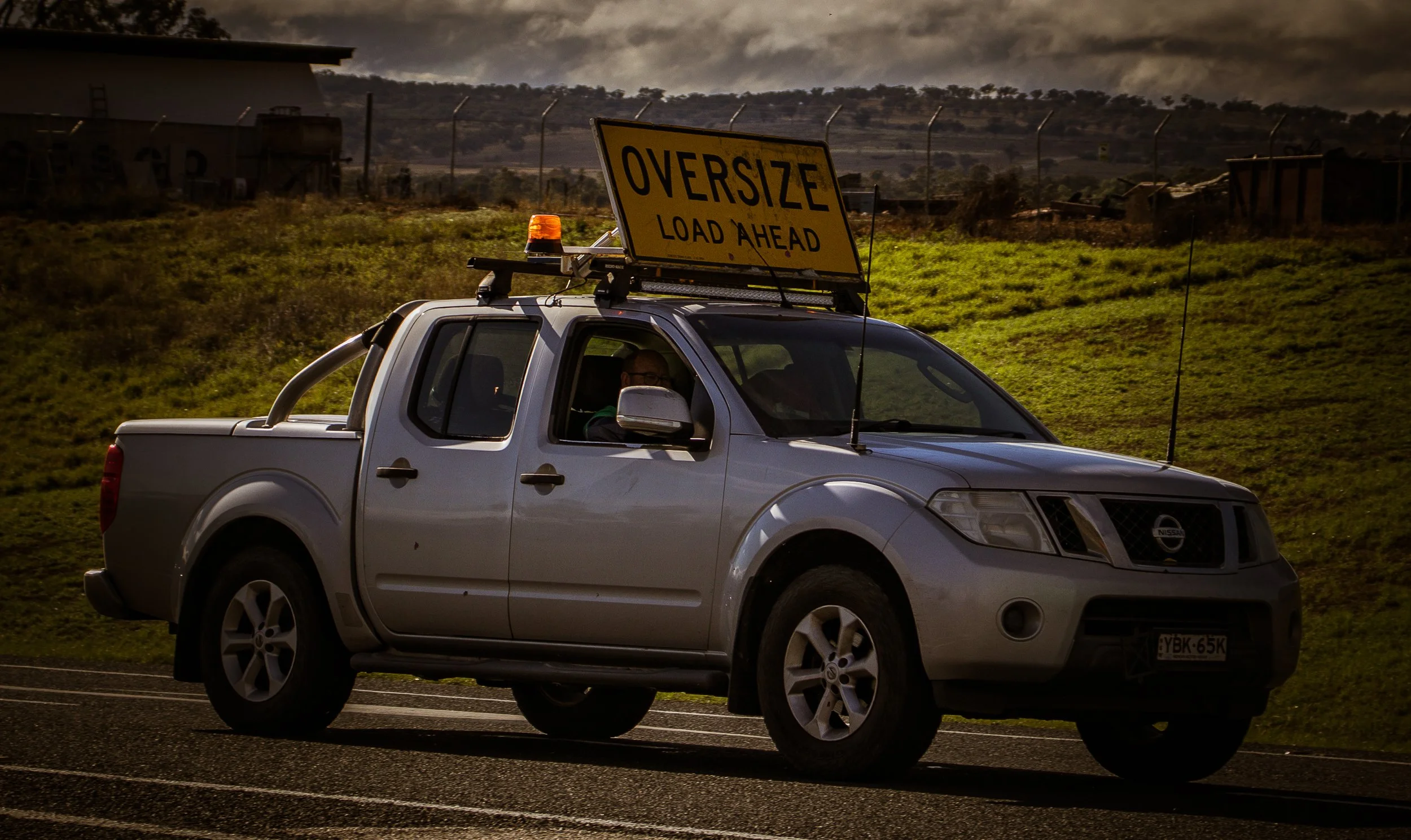 White Nissan pickup truck carrying an illuminated “OVERSIZE LOAD AHEAD” warning sign on a road.