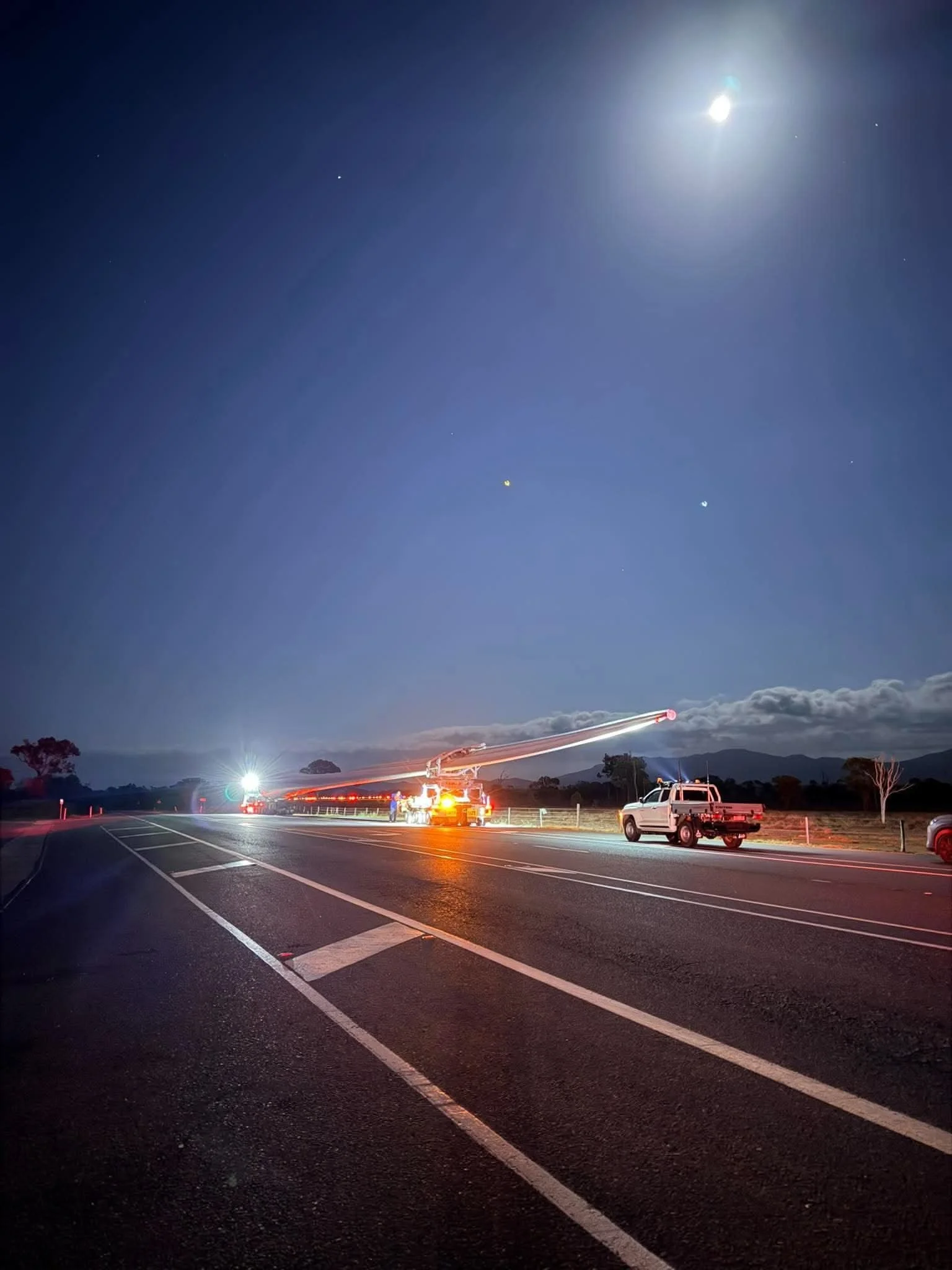 Nighttime roadside scene with a brightly lit emergency truck, extended boom, pickup truck, highway markings, mountains, clouds, and a moonlit sky