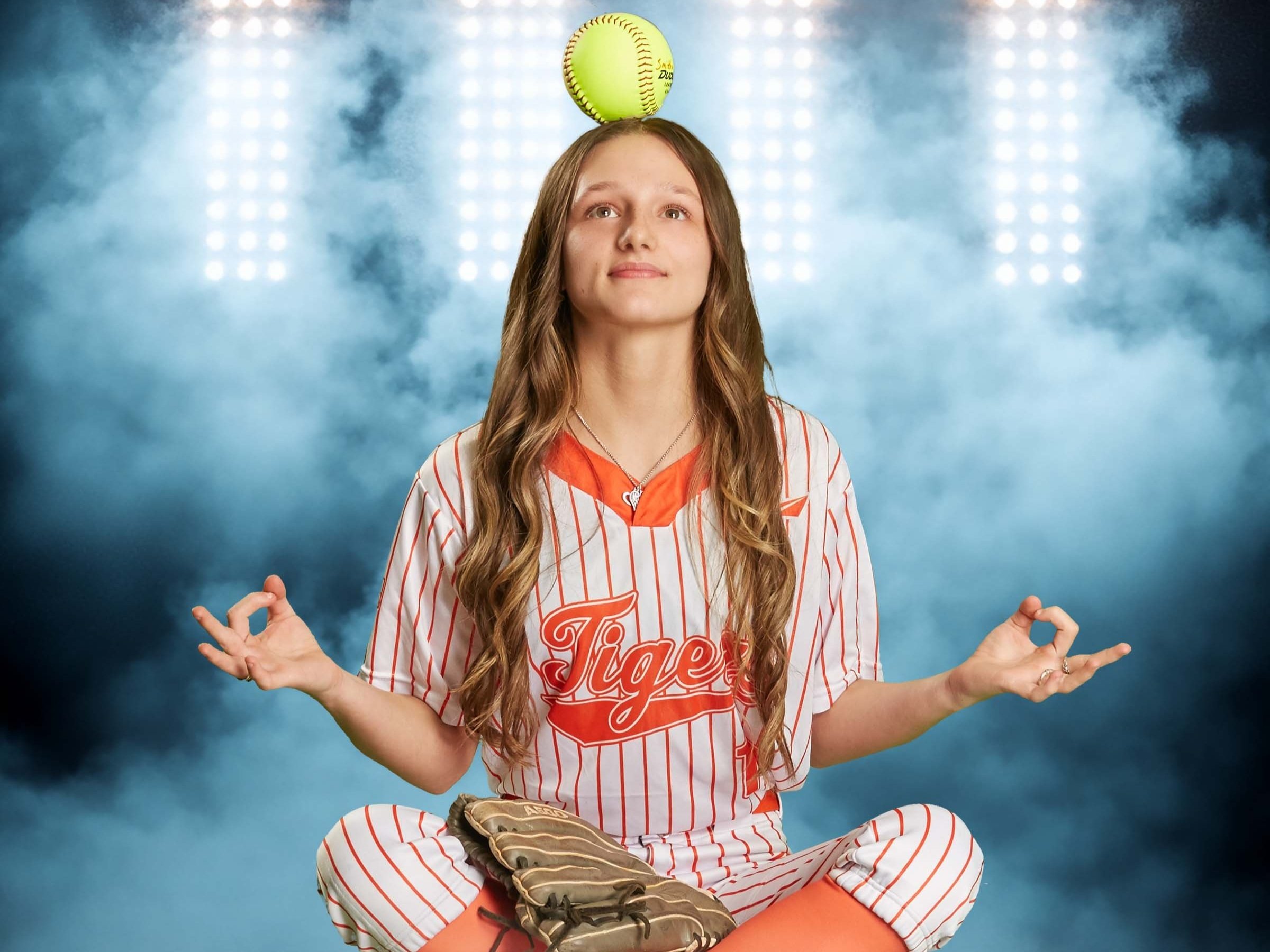 A young woman in a baseball uniform sitting cross-legged, with a softball balanced on her head, in a meditative pose, against a smoky blue background.