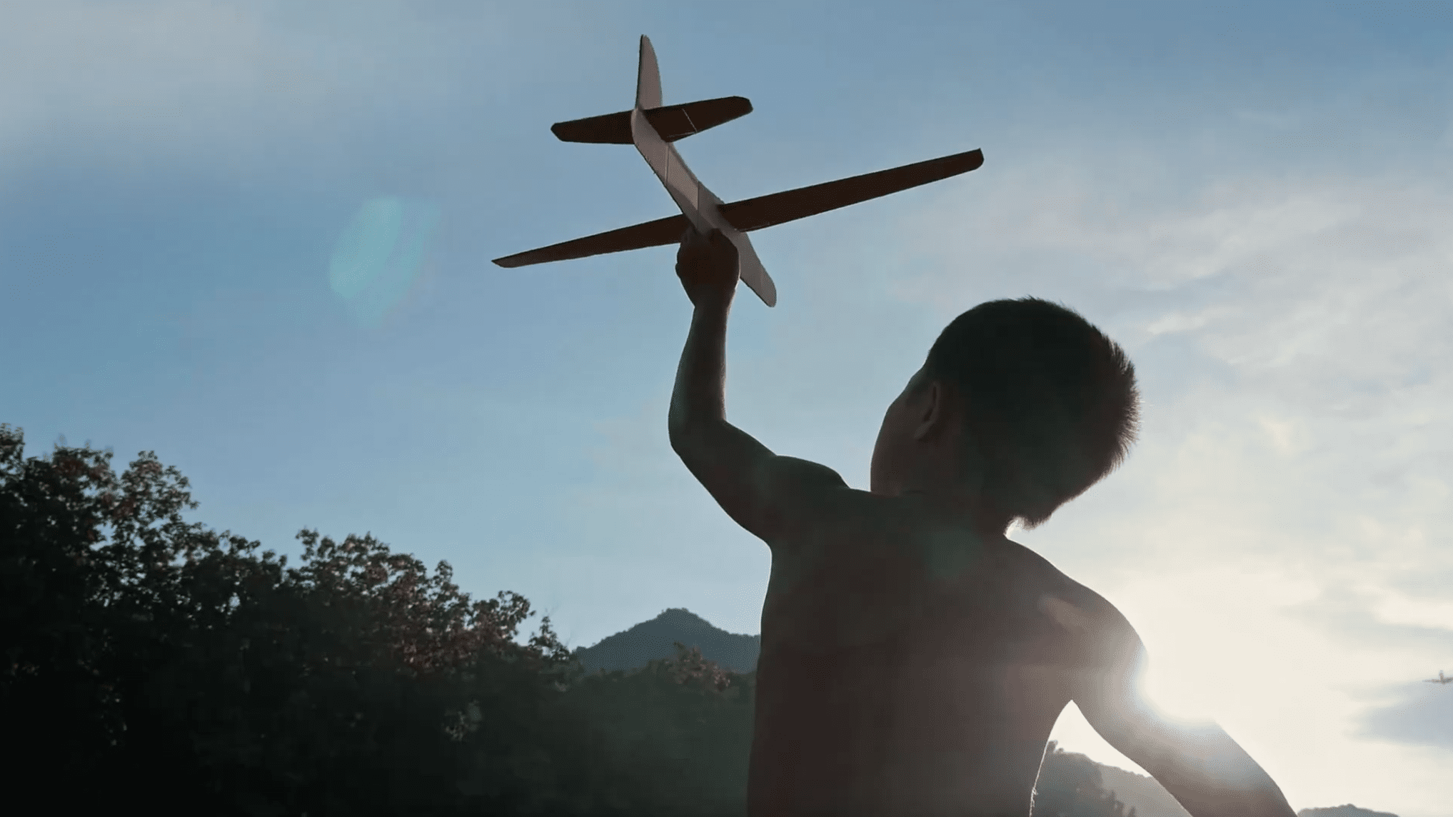 Silhouetted child holding a model airplane overhead at sunset with trees and mountains in the background