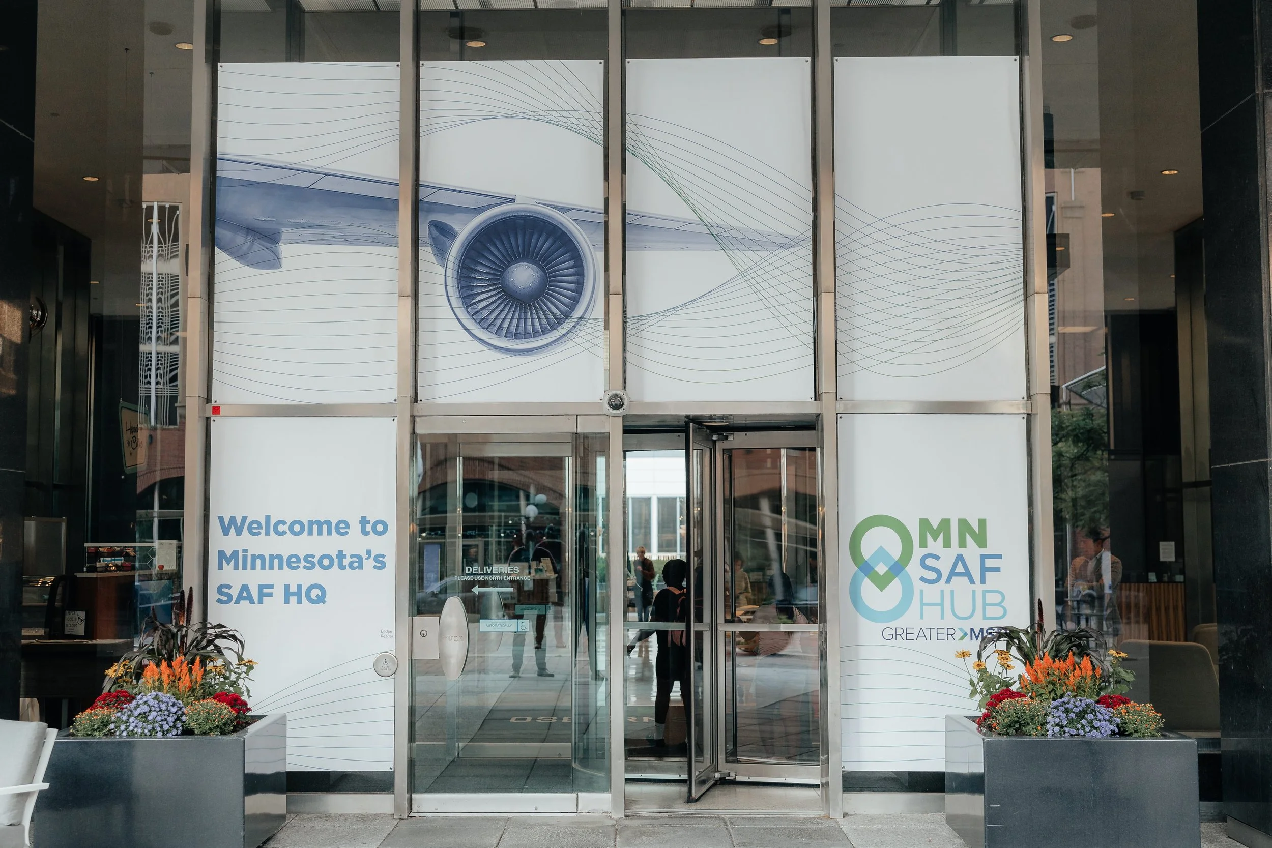 Entrance to Minnesota's SAF Hub headquarters with glass doors, aviation turbine graphic, signage, and flower planters.