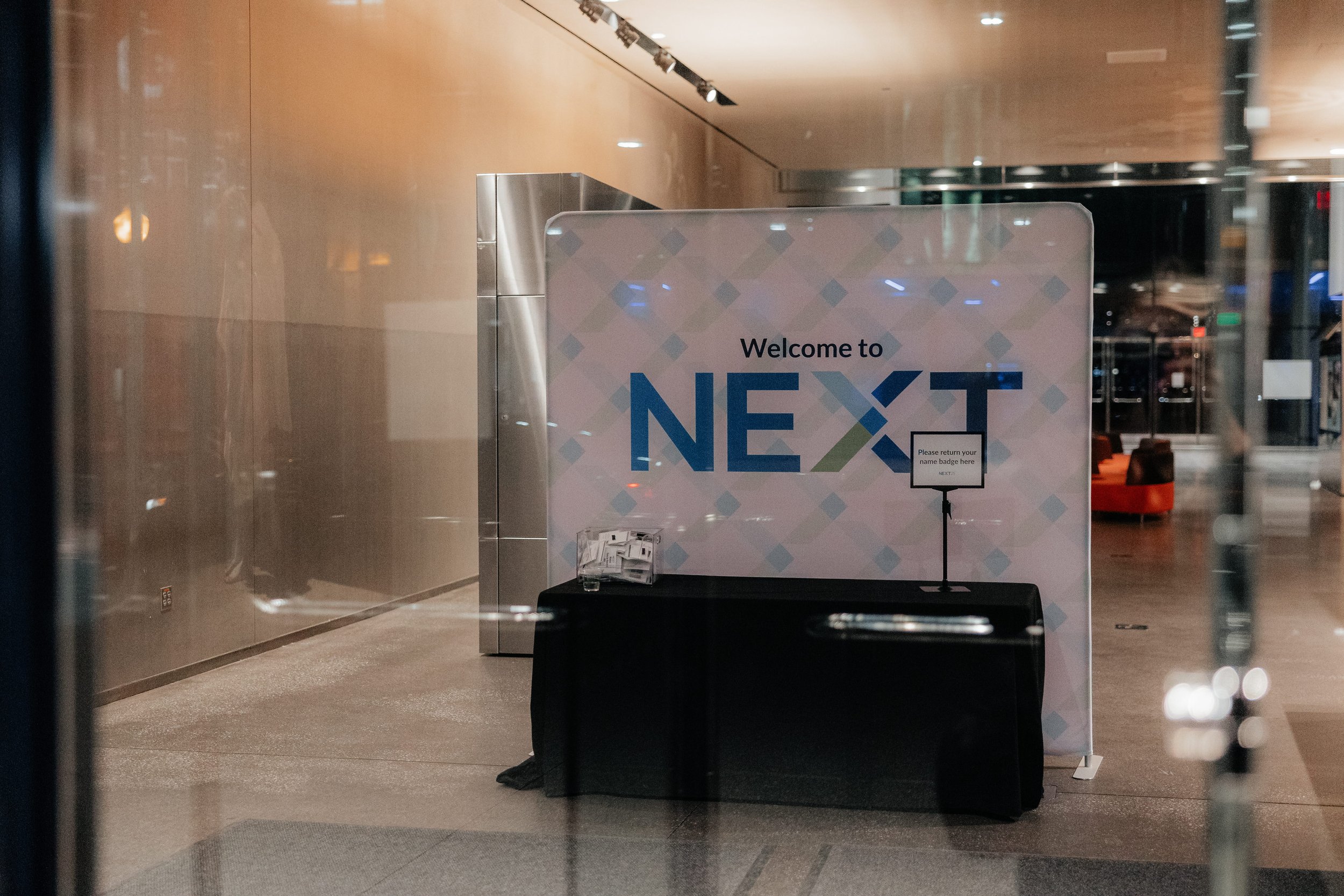 Event welcome table with a NEXT-branded backdrop, name badge return sign, and registration materials in a modern lobby.