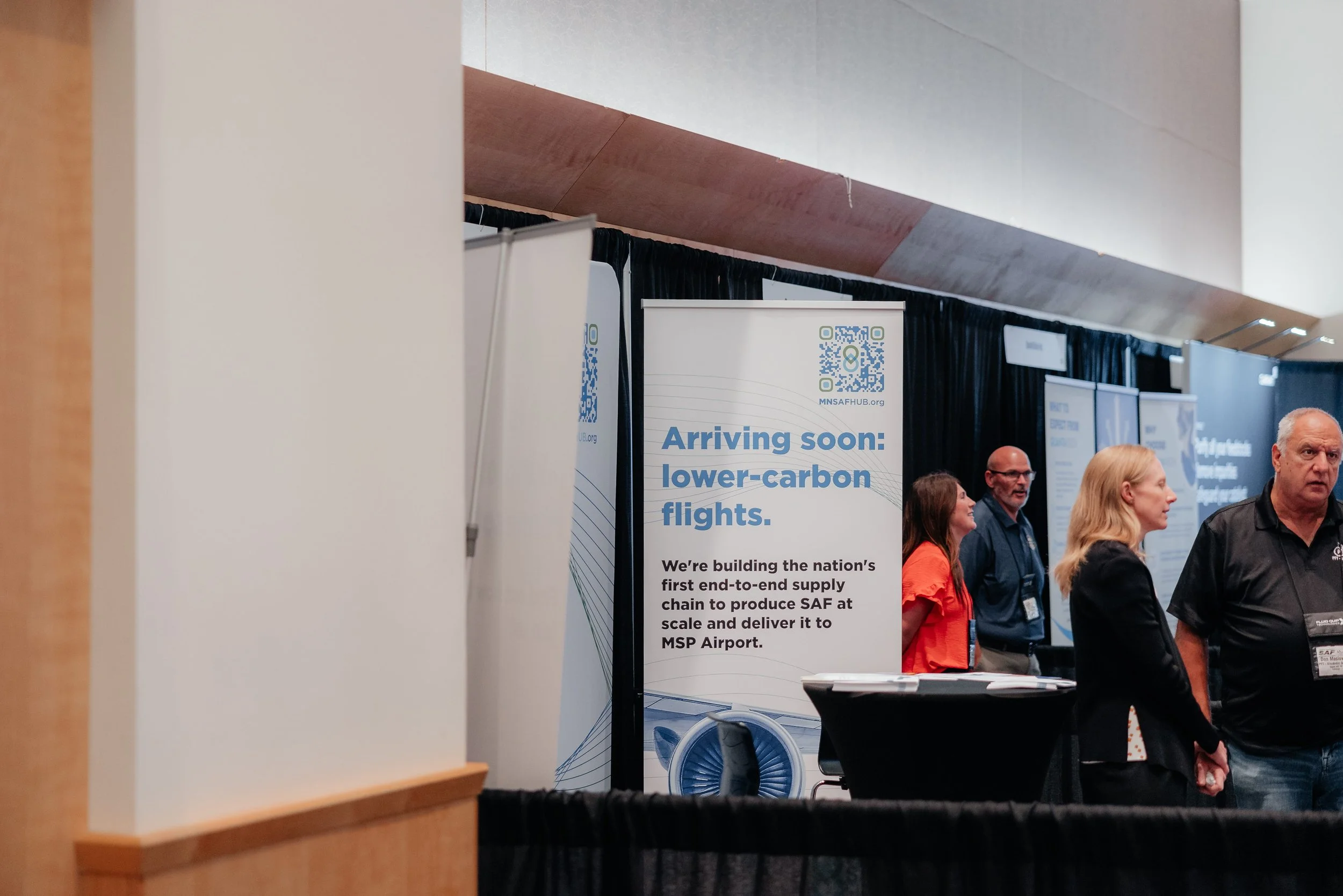 People network at an aviation sustainability expo beside a banner promoting lower-carbon flights and sustainable aviation fuel at MSP Airport.