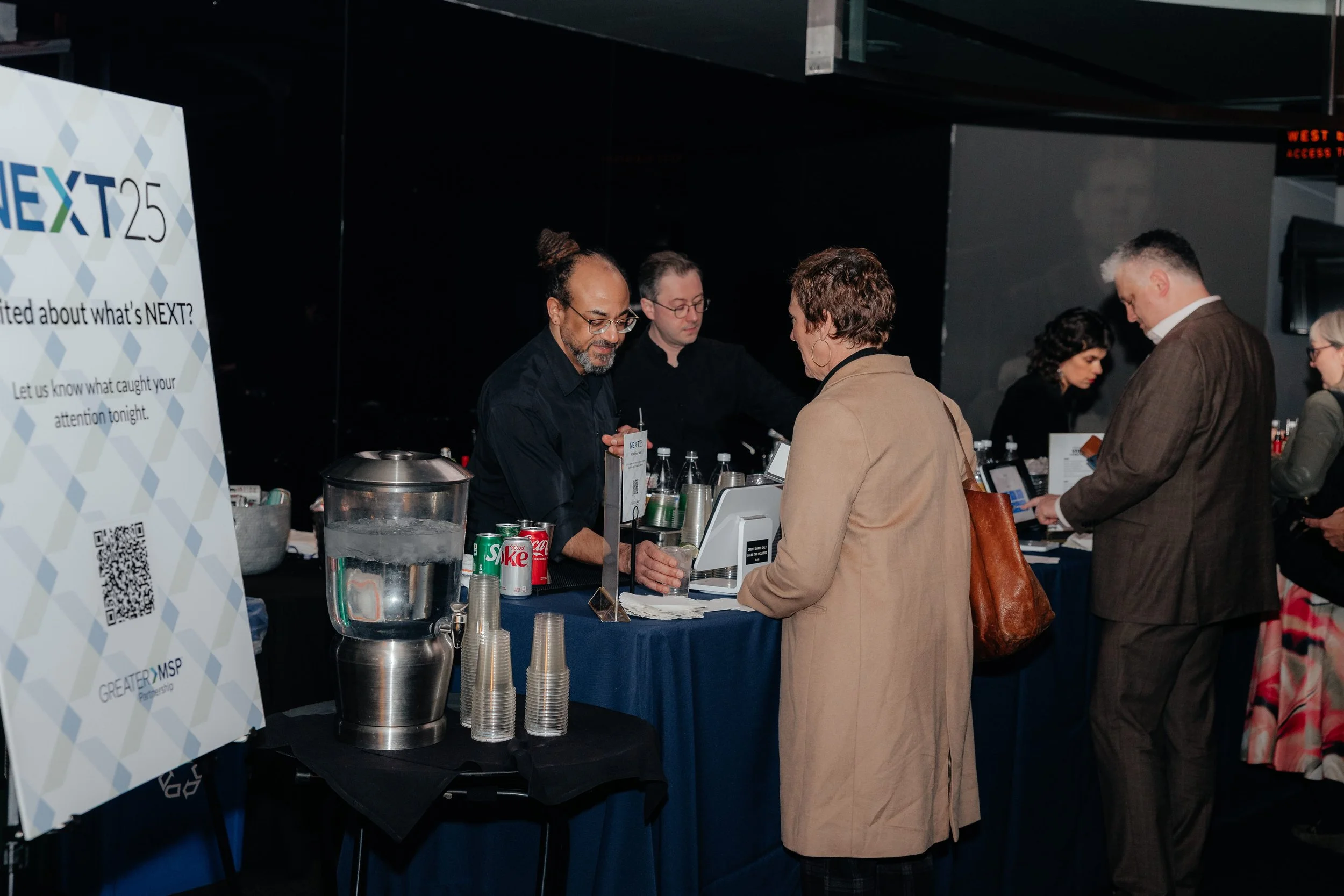 Guests mingle at a NEXT25 event beverage station with drinks, ice, and stacked cups.