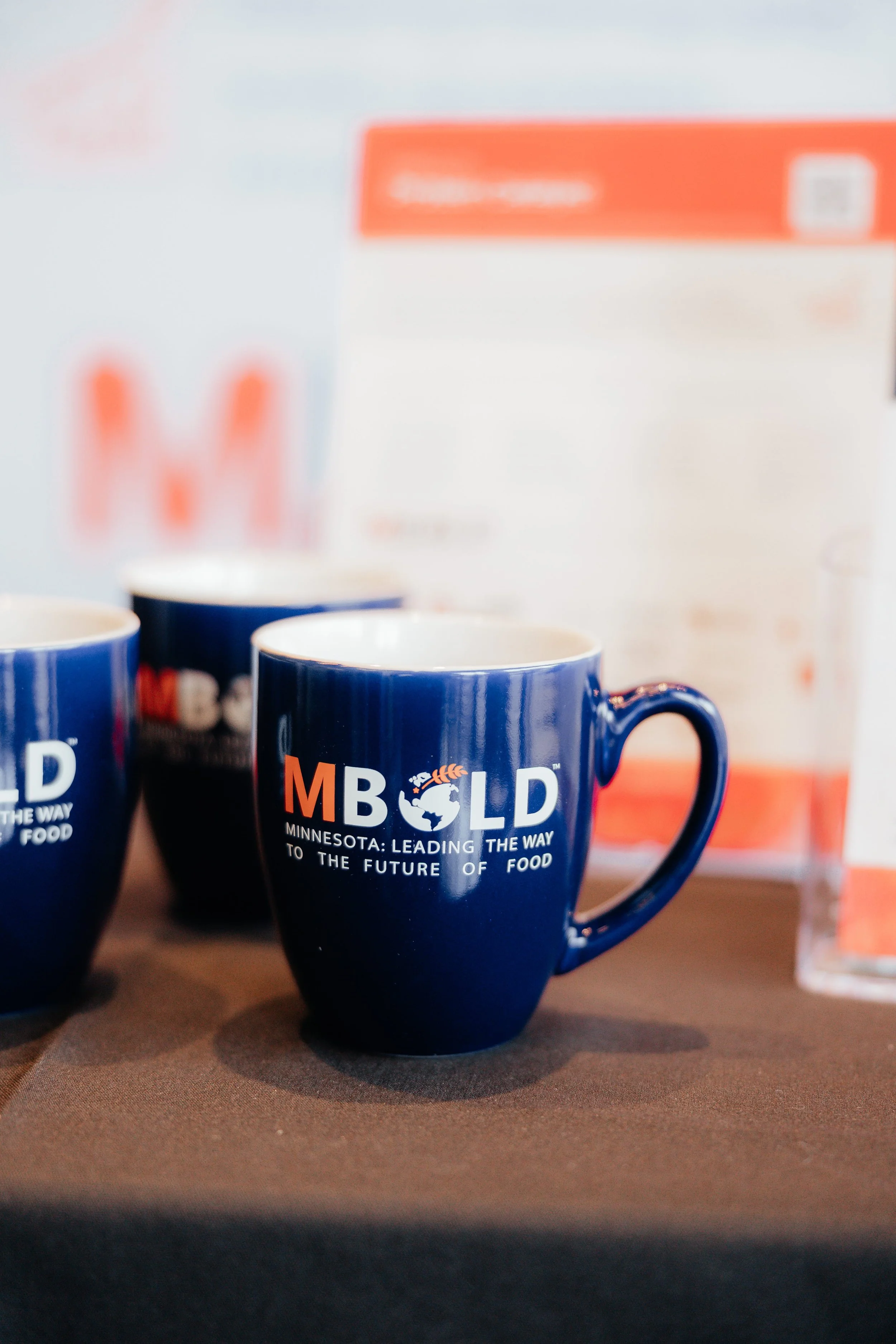 Blue “MPLD” coffee mug with a globe logo and “Minnesota: Leading the Way to the Future of Food” text on a wooden table.