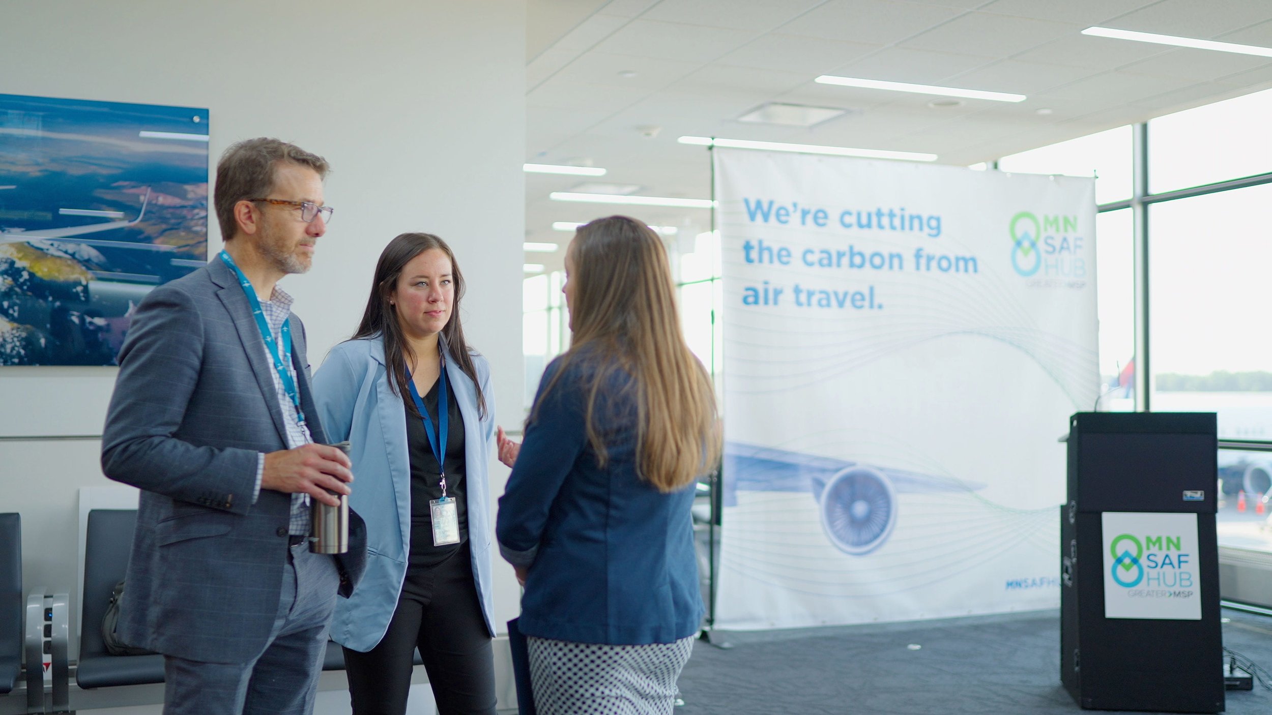 Three professionals talk beside an airport sustainability display reading “We’re cutting the carbon from air travel.”