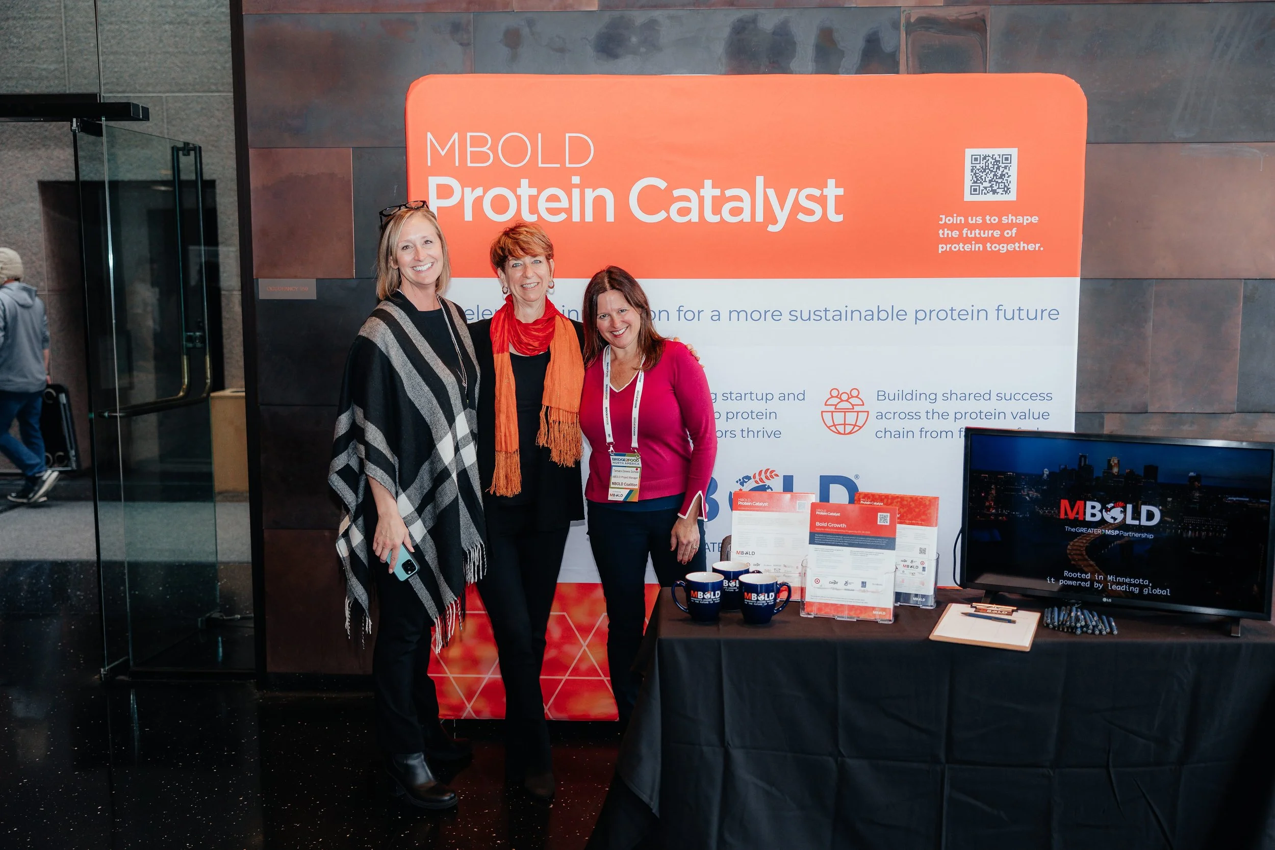 Three women pose at an MBOLD Protein Catalyst event booth with promotional materials, branded mugs, and a monitor.