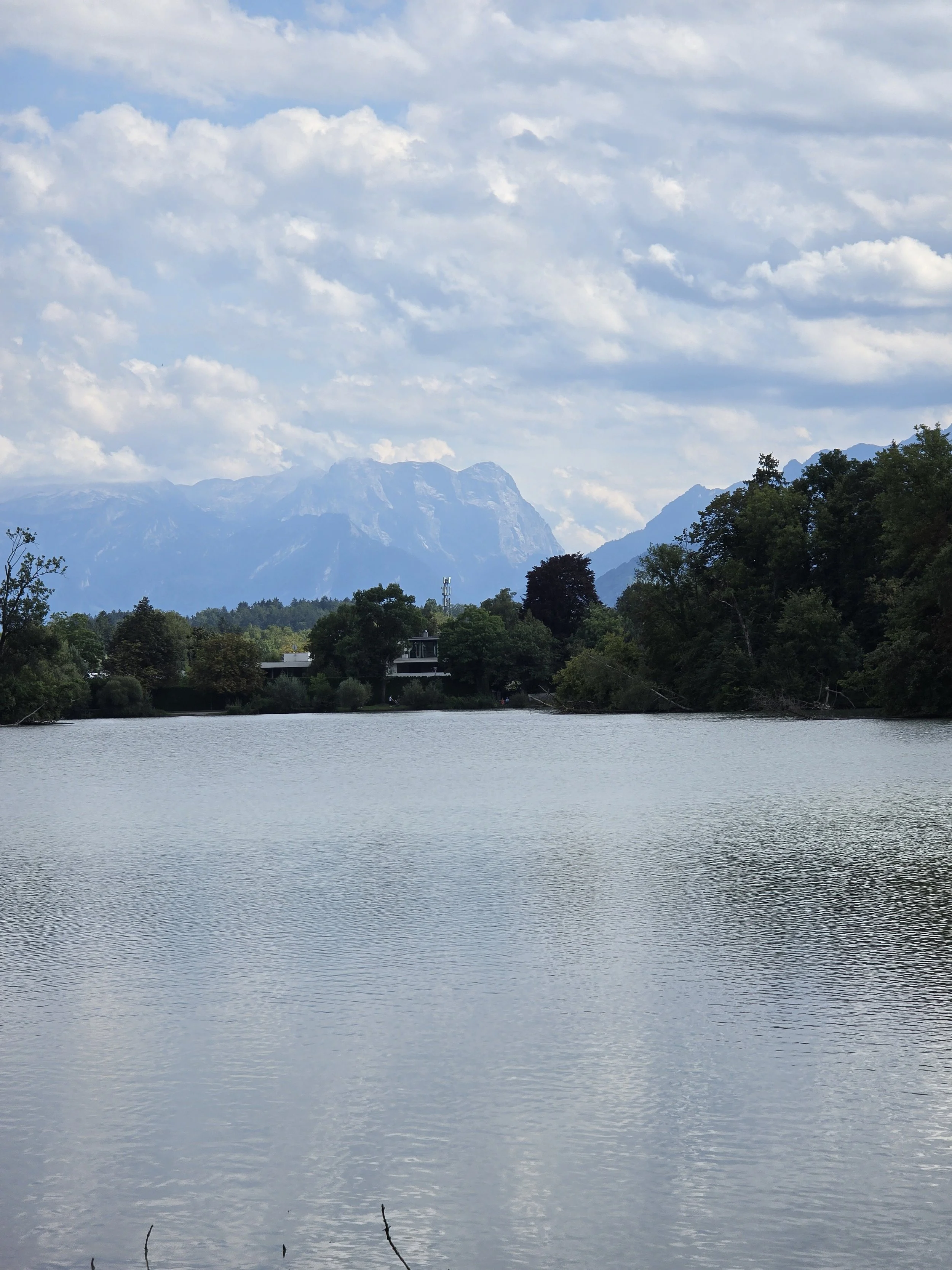 Lake with forested shoreline and dramatic mountains beneath a cloudy blue sky