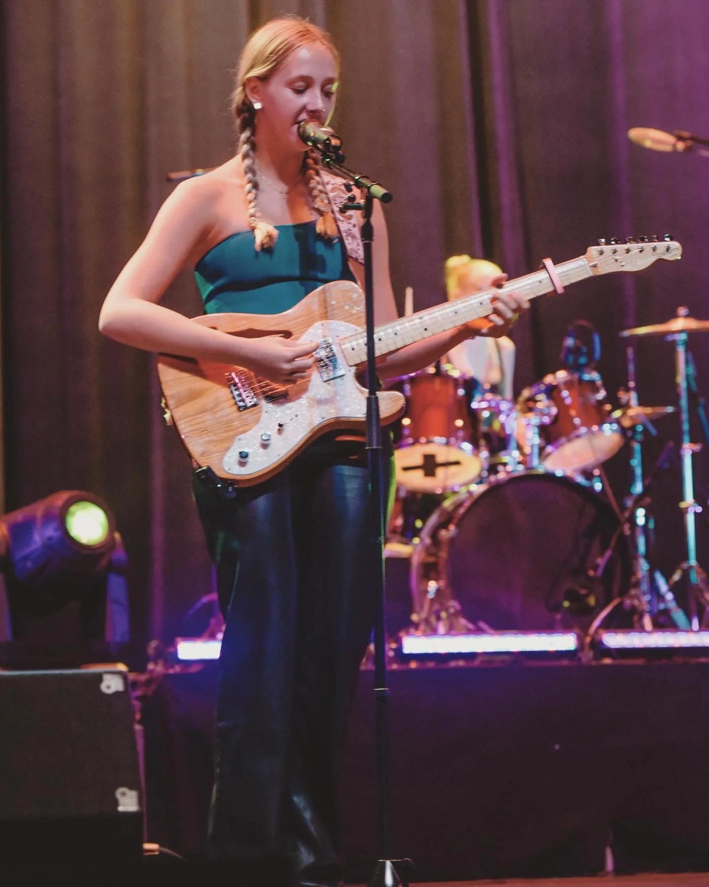 Female musician singing into a microphone while playing electric bass on stage, with a drummer and colorful concert lights behind her