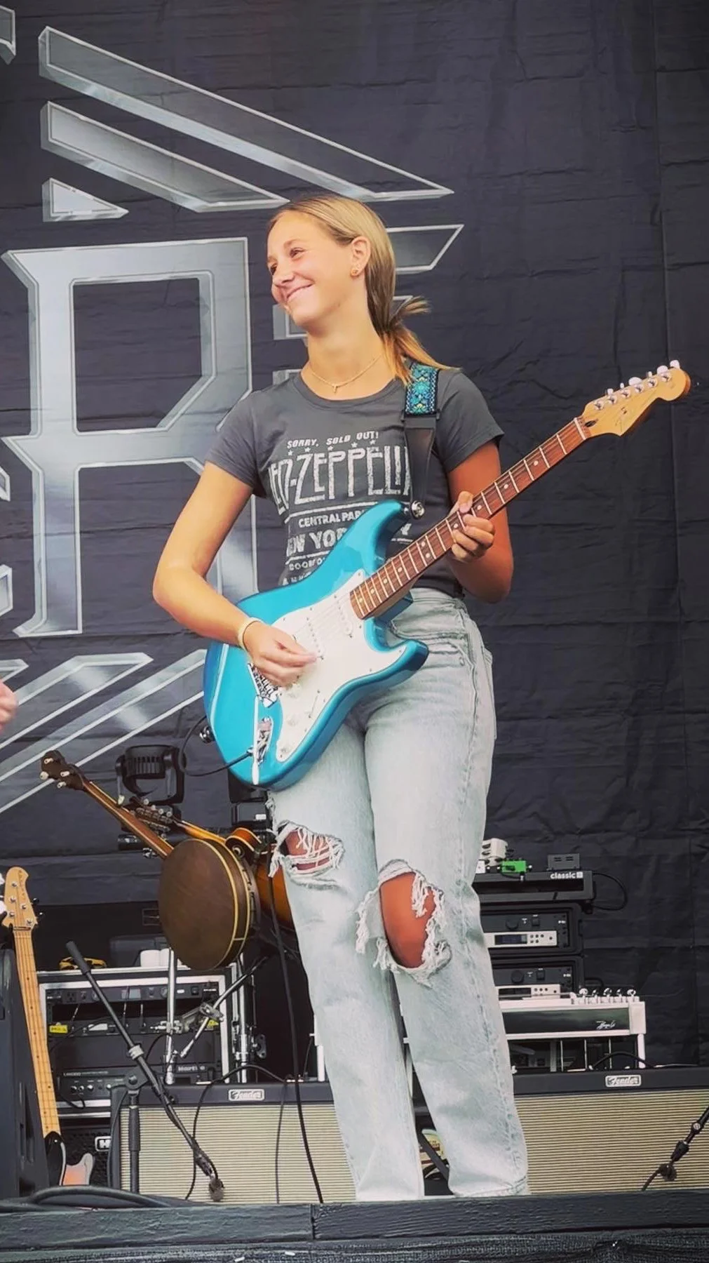 Musician playing a turquoise electric guitar onstage in front of amplifiers.