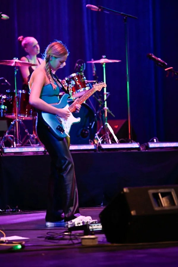 Female guitarist performing onstage with a drummer under purple and blue concert lights