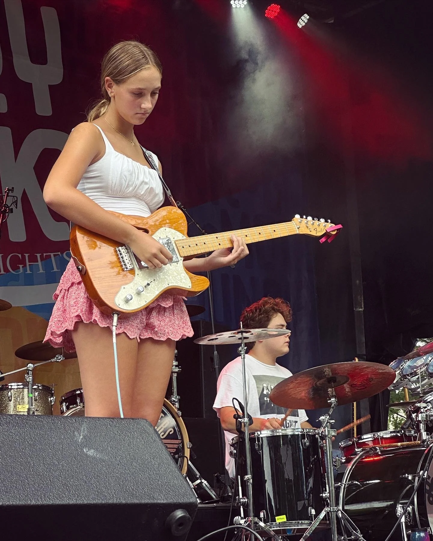 Young female guitarist performing onstage beside a drummer under red and white concert lights