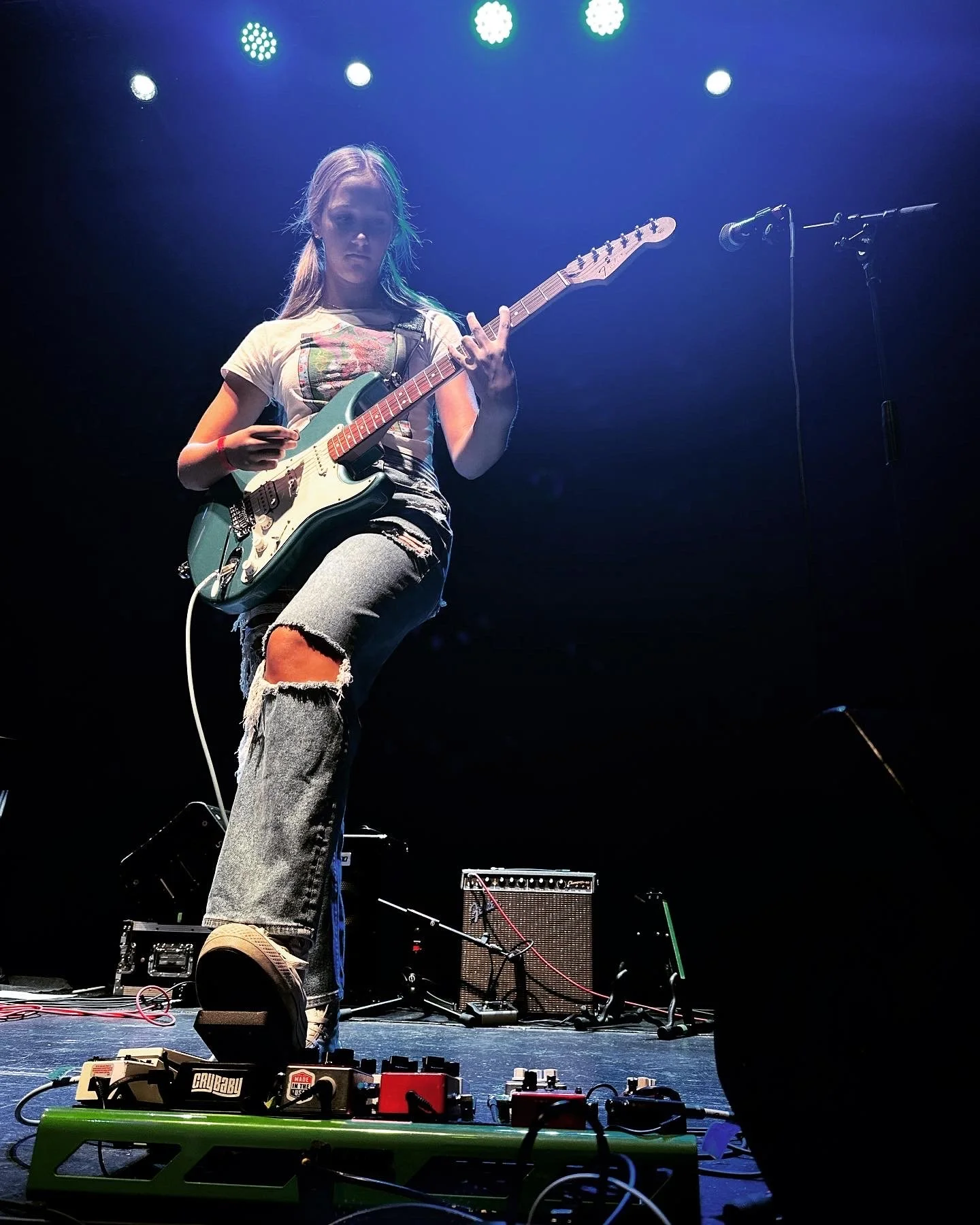 Guitarist performing onstage under blue lights with an electric guitar and pedalboard