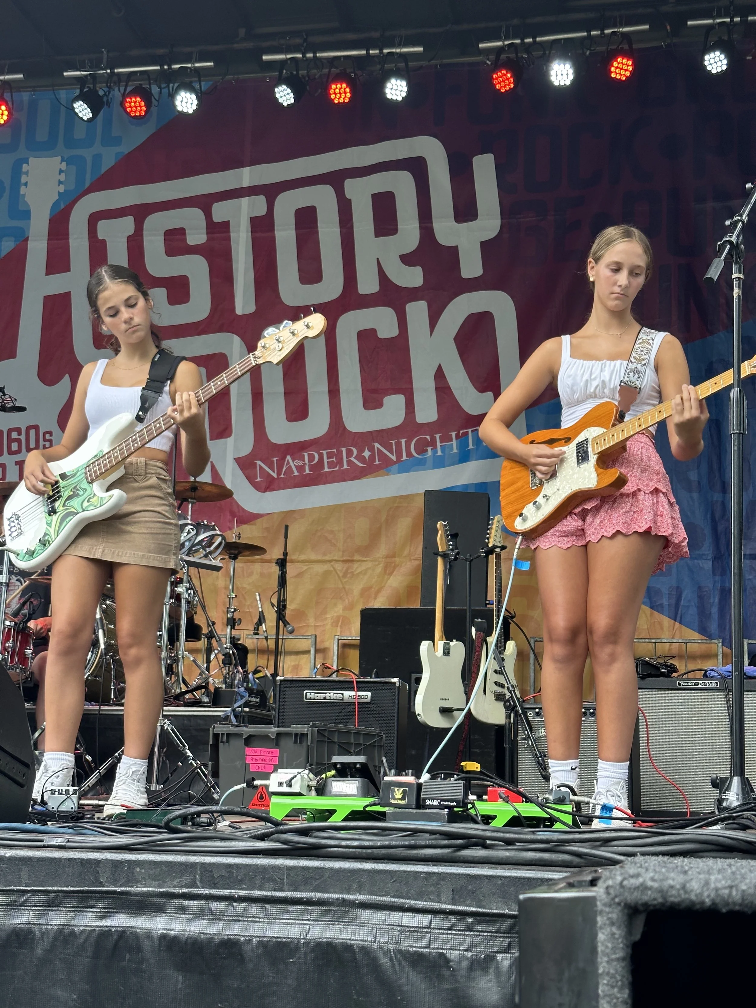 Two female musicians perform onstage, playing electric bass and guitar beneath colorful concert lights.