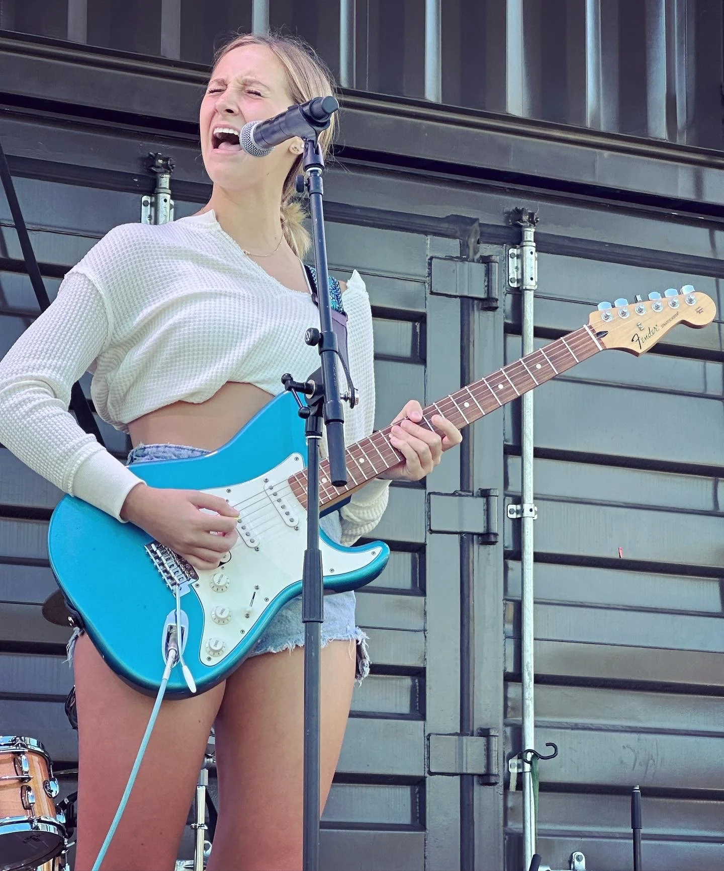 Musician singing into a microphone while playing a blue electric guitar onstage.