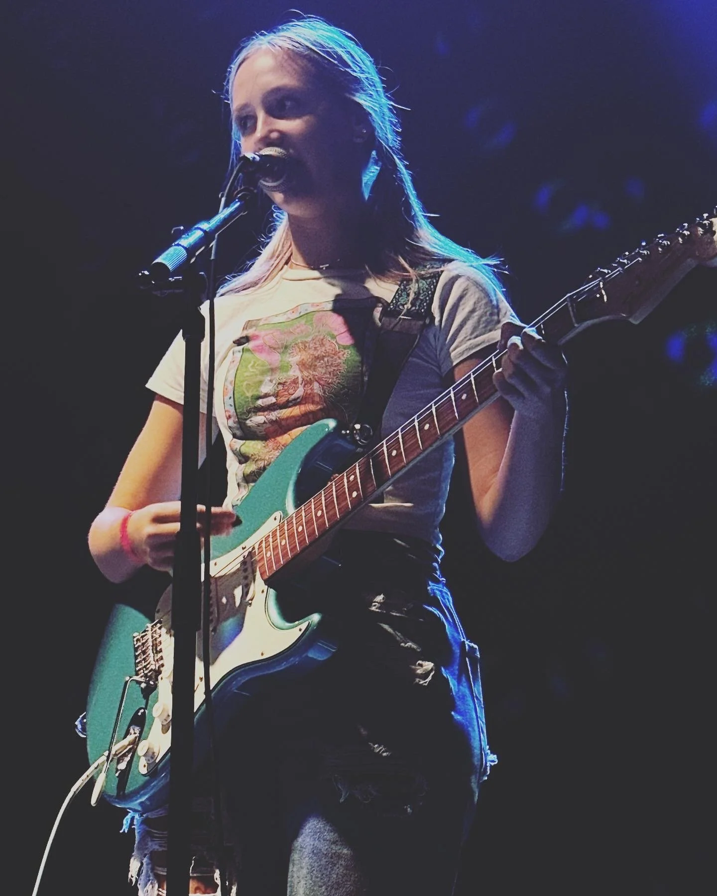 Female guitarist singing into a microphone during a live concert under blue stage lighting