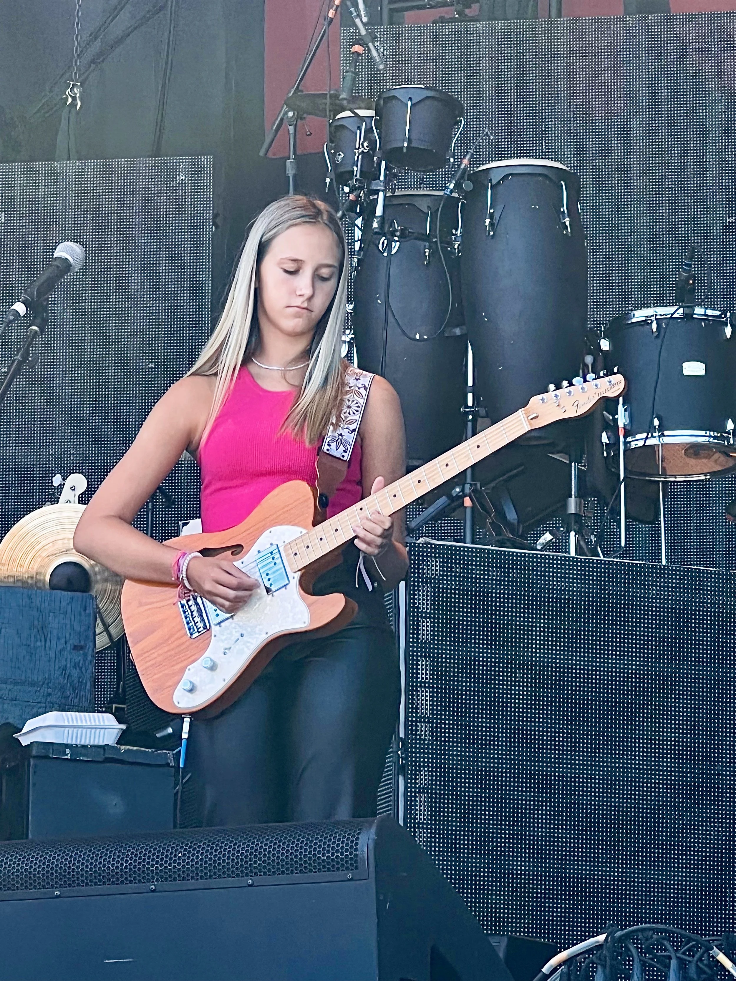 Young woman in a pink top playing electric guitar on a concert stage with a microphone and drum kit behind her