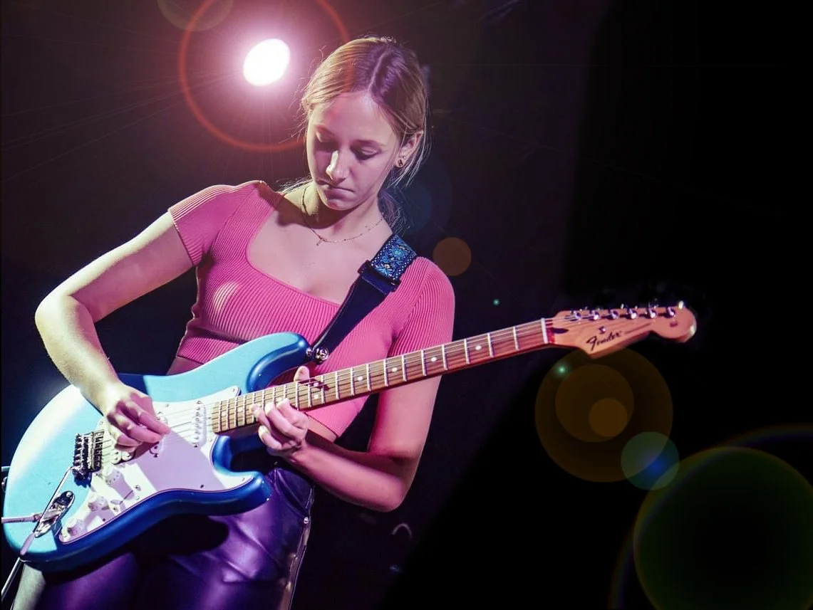 Female guitarist playing a blue electric guitar under colorful stage lights