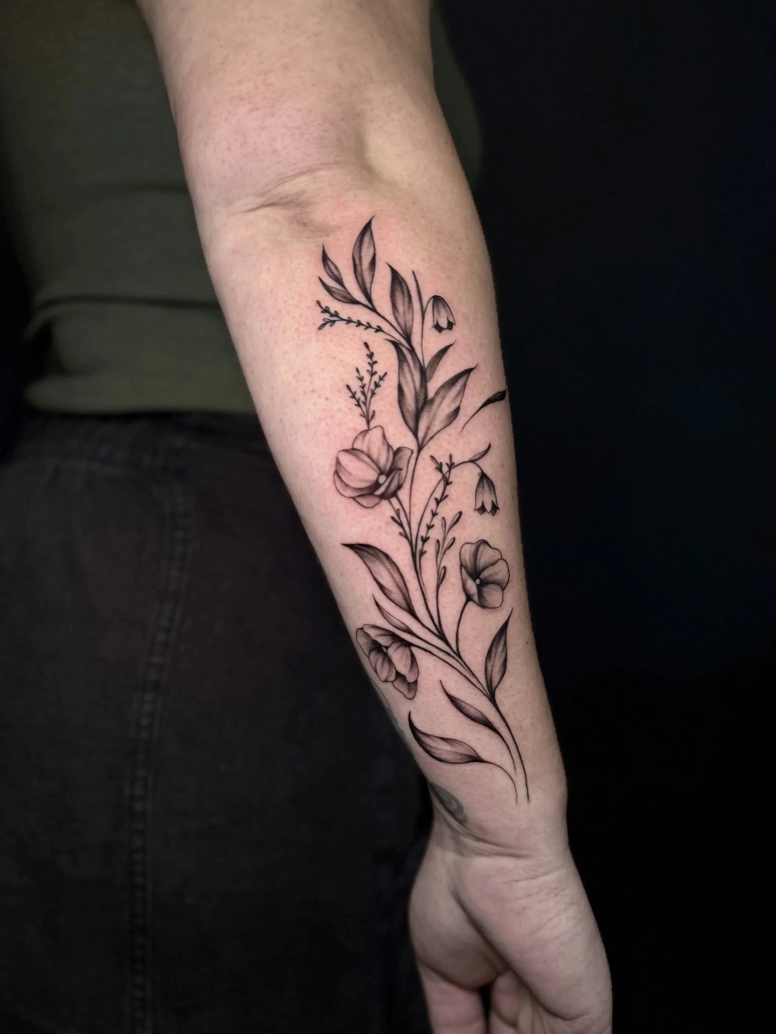 Close-up of a forearm with a detailed black-and-gray floral tattoo featuring flowers, leaves, and stems.