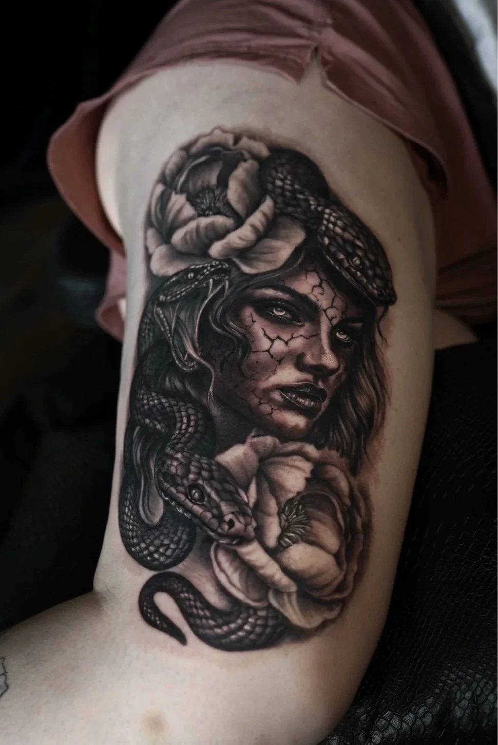 Detailed black-and-gray tattoo of a cracked-faced woman with snakes and flowers on an upper arm